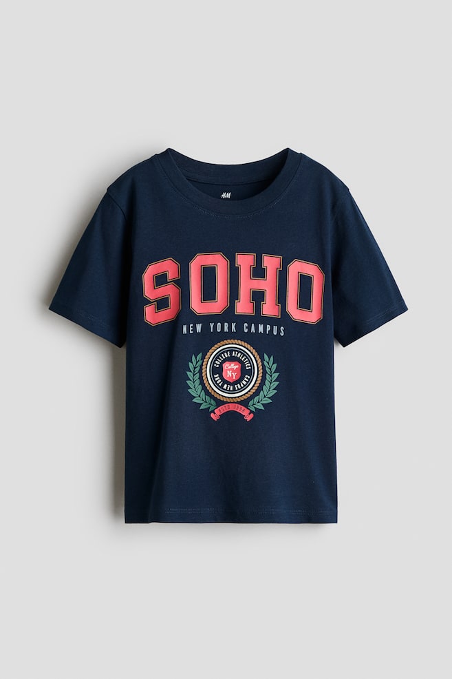 A navy blue cotton jersey T-shirt is shown from the front, featuring a round ribbed neckline and short sleeves with gently dropped shoulders. The chest displays a printed collegiate graphic with "SOHO" in large pink block letters above the text "NEW YORK CAMPUS" and a circular athletic crest. - 1