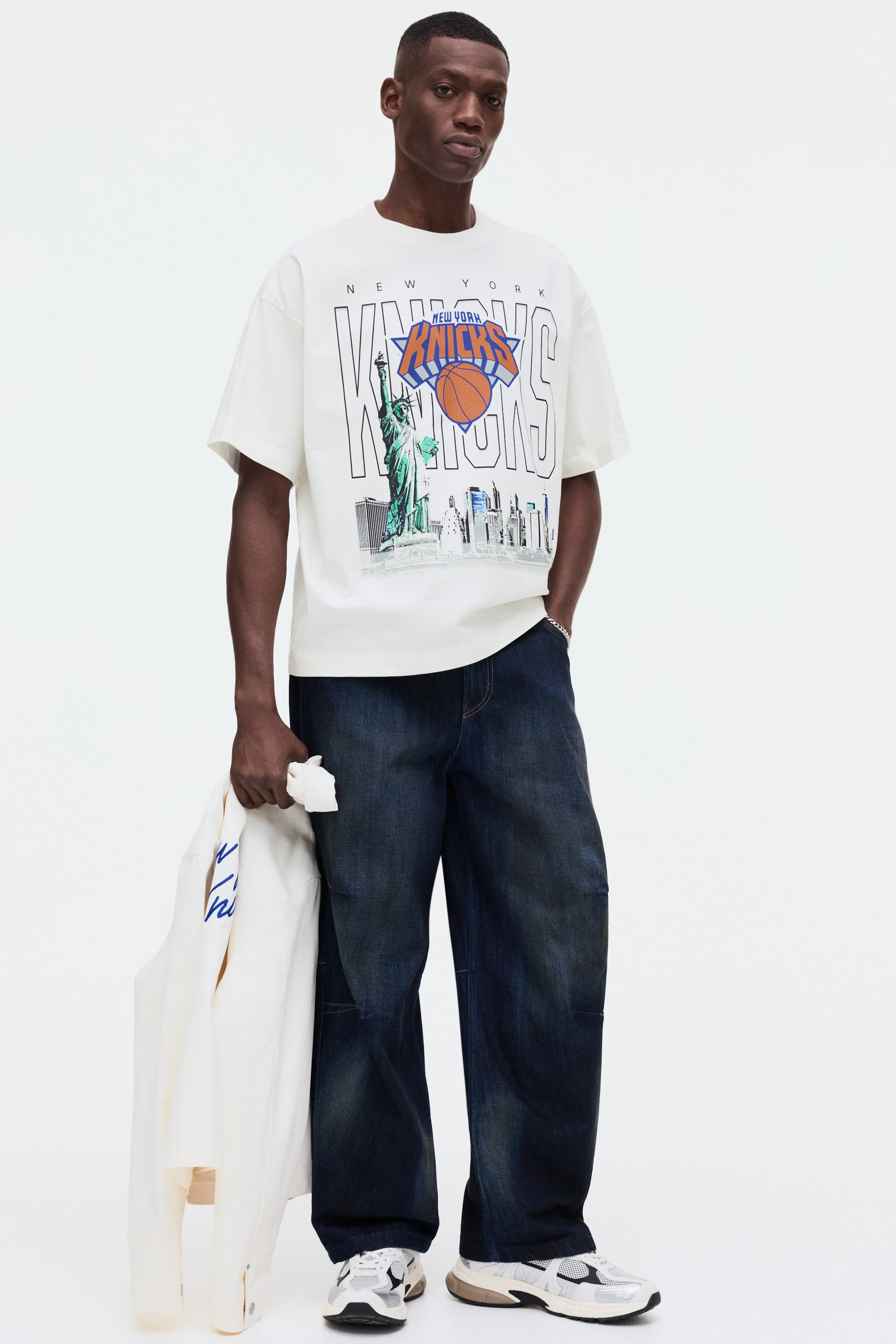 Visa större bild: Den vita t-shirten har ett stort grafiskt tryck på framsidan med texten "New York Knicks", en orange basketboll, Frihetsgudinnan och en stadssilhuett. Den har en avslappnad passform med rund hals och korta ärmar.