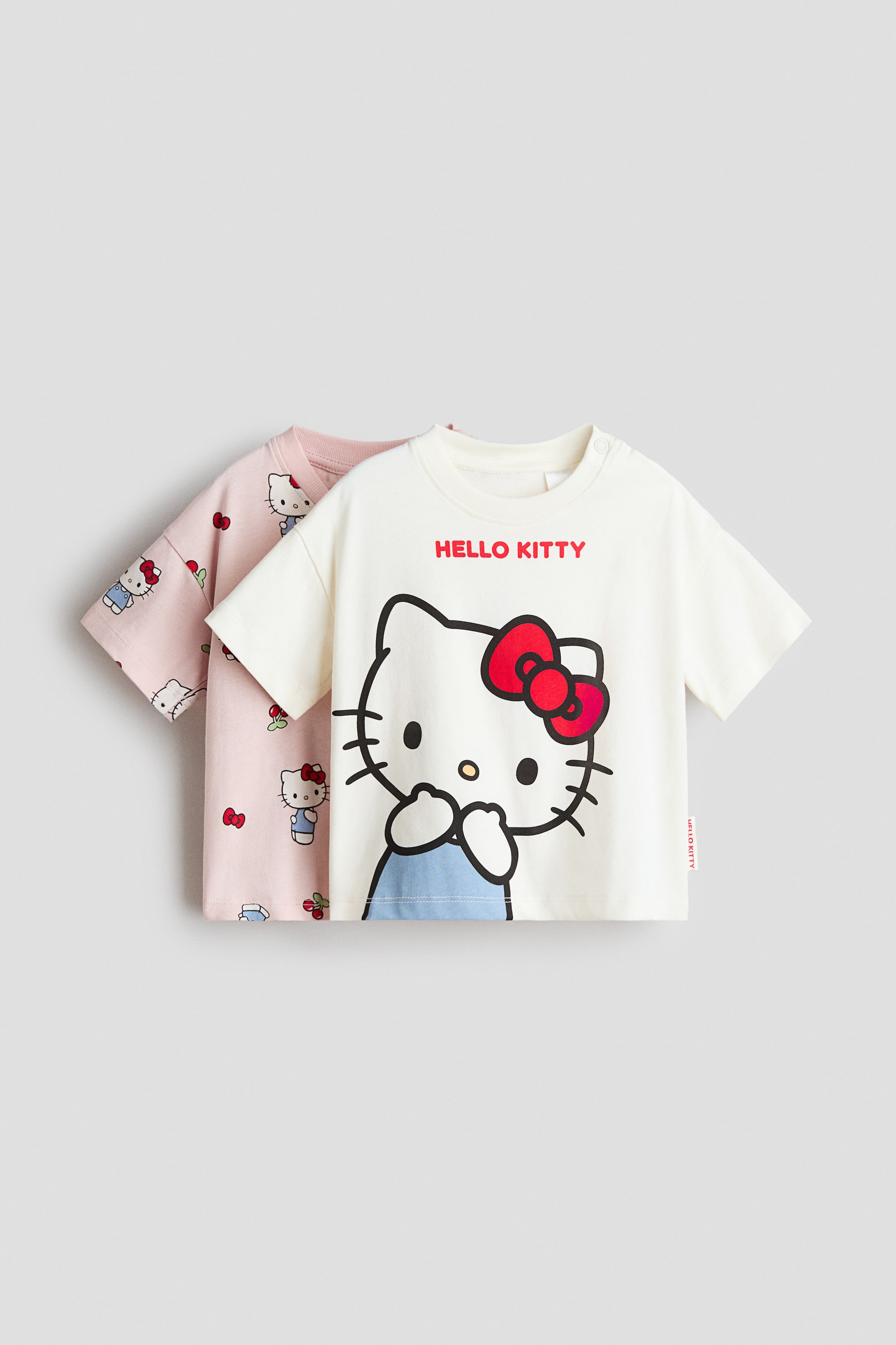 View larger image: Presented is a 2-pack of printed cotton T-shirts, featuring an off‑white T-shirt at the front and a pink T-shirt layered behind it. The front off‑white T-shirt displays a large Hello Kitty graphic with a red bow and light‑blue overalls, along with the "HELLO KITTY" text in red and snap buttons on the left shoulder, while the pink T-shirt features an all‑over print of smaller Hello Kitty faces, red bows and cherries.