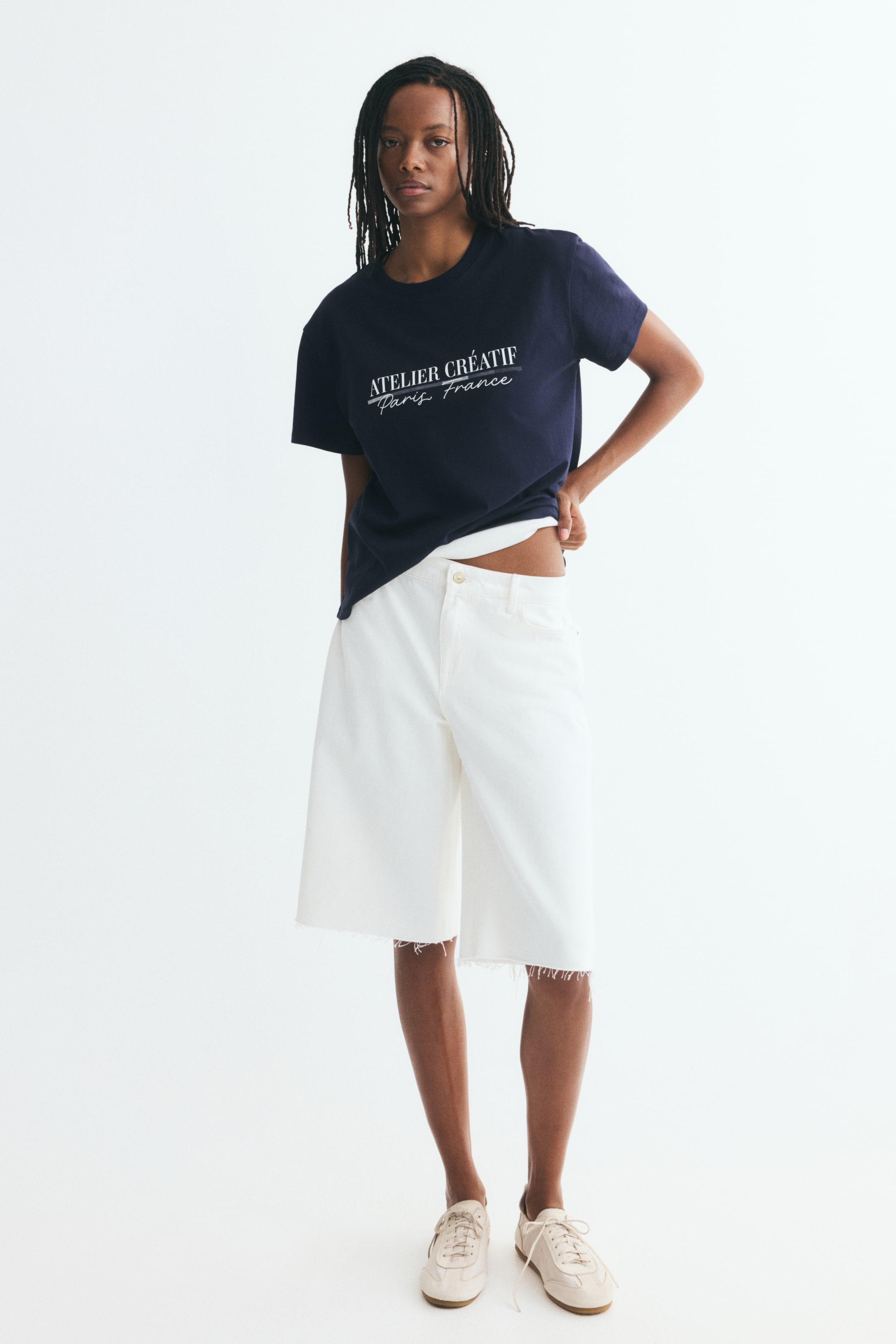 Xem ảnh lớn hơn: Một chiếc áo thun màu xanh navy, dáng rộng, làm từ cotton jersey mềm mại đang được hiển thị ở góc chụp chính diện, với cổ tròn viền bo gân, vai trễ và họa tiết chữ in màu trắng ghi "ATELIER CR�ATIF Paris France" ngang ngực. Áo rủ nhẹ theo dáng cơ thể và được phối với phần gấu áo hơi kéo cao một bên bên trên quần short denim trắng.