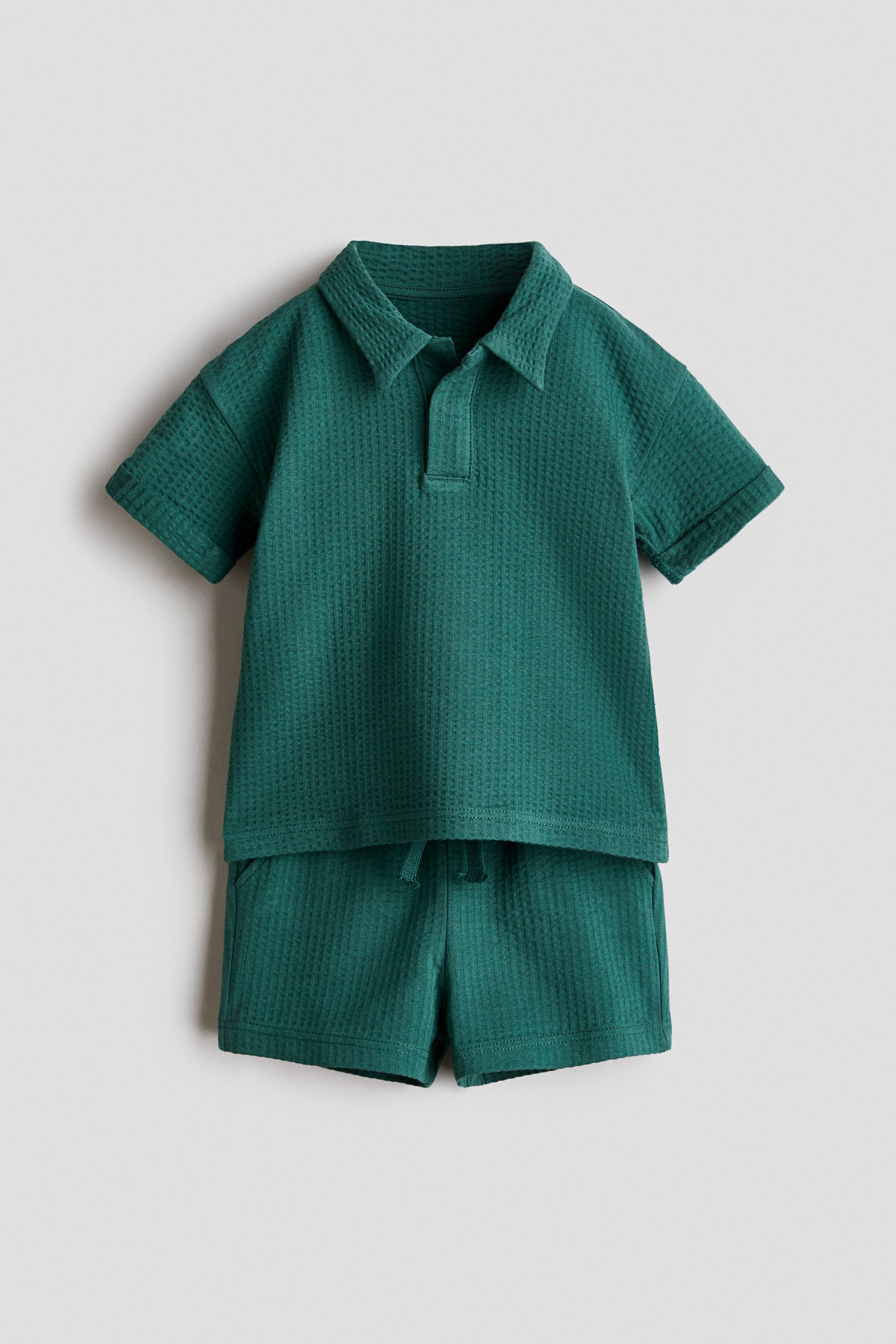 Agrandir l'image: Un ensemble deux pièces en maille gaufrée bleu canard profond est présenté, comprenant un polo à manches courtes avec un col classique et une patte de boutonnage courte, ainsi que des poignets retroussés. Le short assorti est doté d'une taille élastique avec un cordon de serrage visible, complétant l'ensemble orienté vers l'avant.