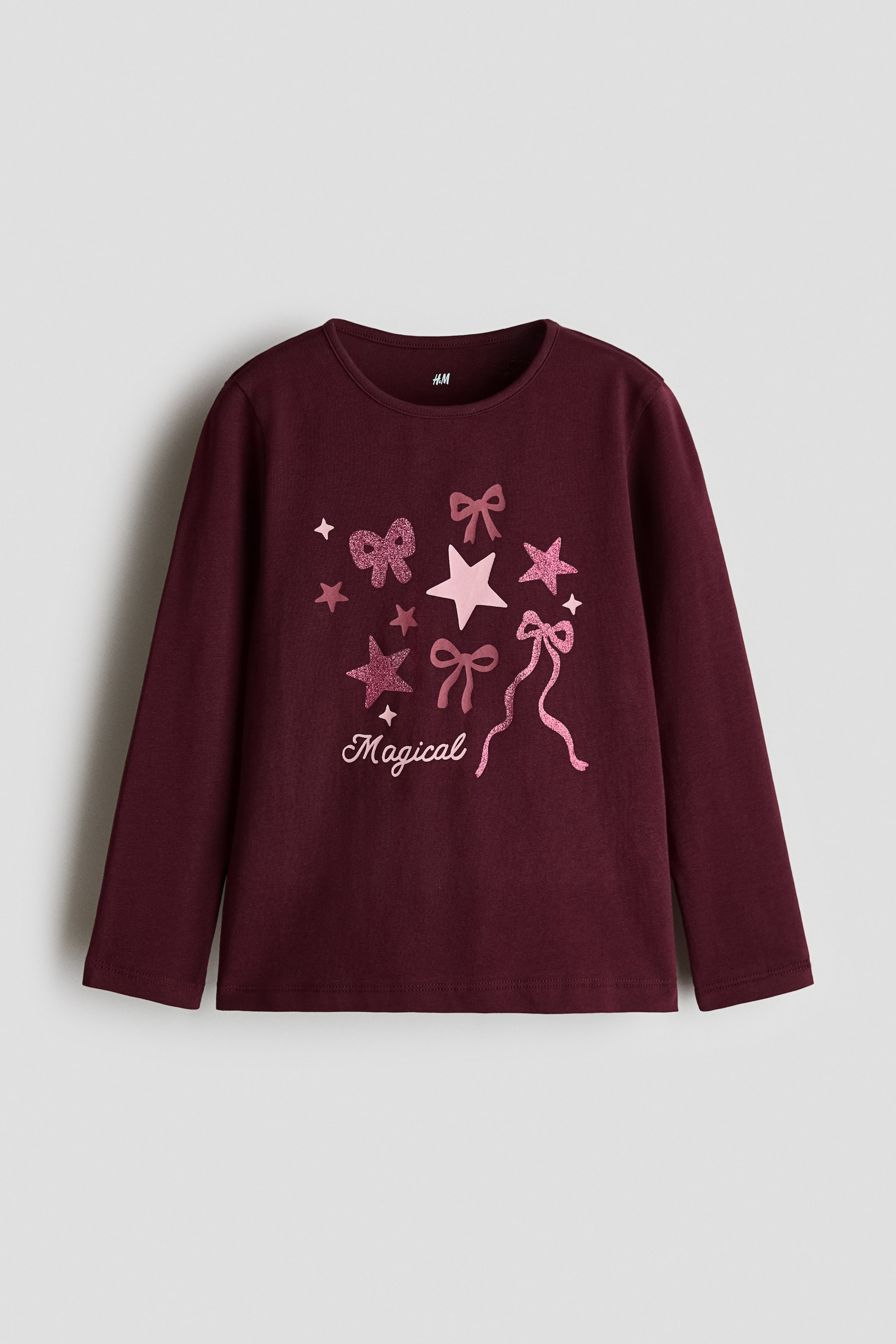 View larger image: A burgundy long-sleeved top is facing forward, featuring a round neckline with a narrow trim. The front displays a pink graphic print with stars, bows, and the script word “Magical” with glitter accents.