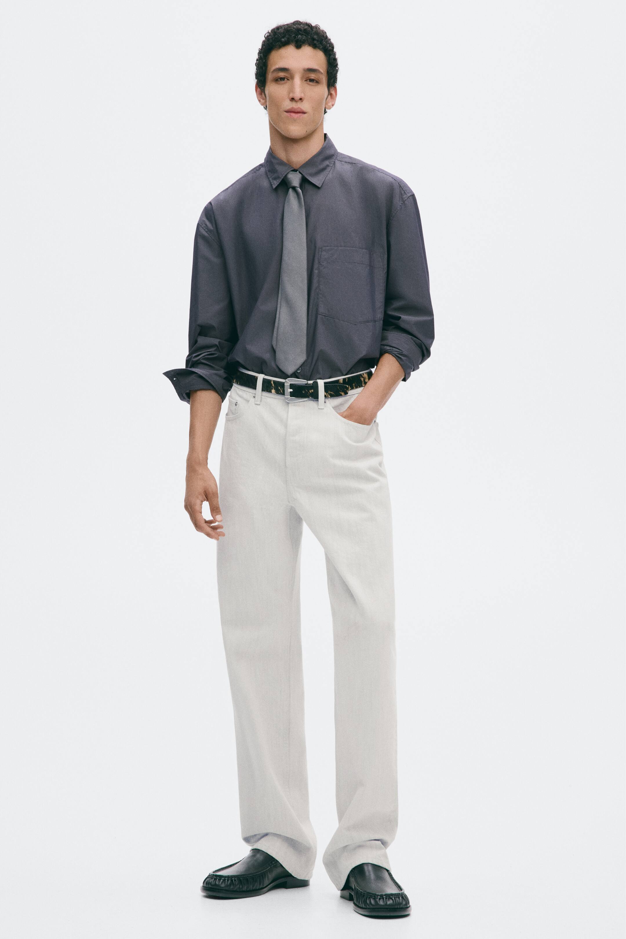 Grösseres Bild ansehen: Offwhite Straight-Leg-Denimjeans mit Regular Fit werden vom Model getragen, das mit einer Hand in der vorderen Tasche steht. Die Jeans sind mit einem gemusterten Gürtel gesichert und mit einem dunkelgrauen Langarmhemd, einer grauen Krawatte und schwarzen Lederschuhen kombiniert.