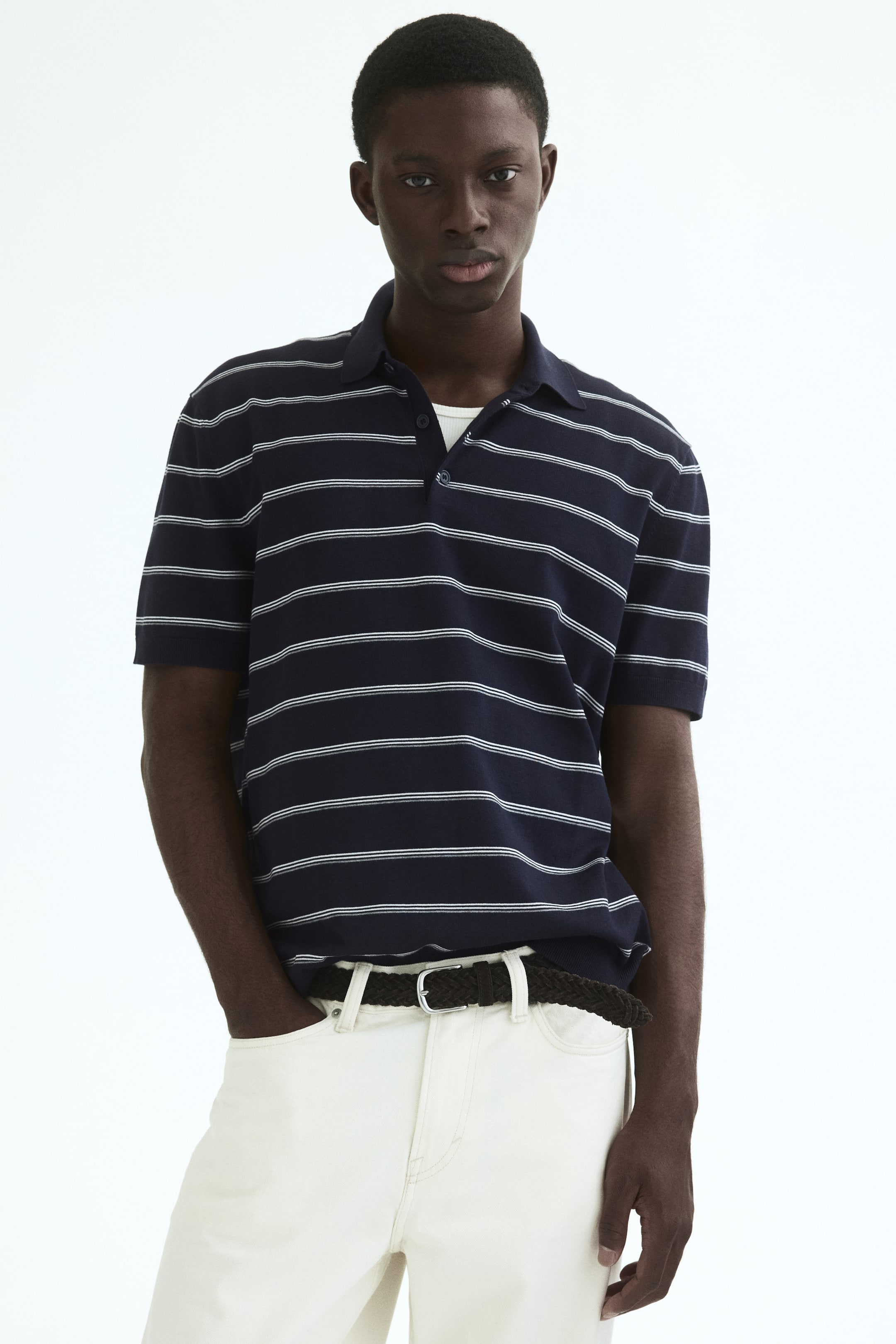 Vezi imaginea mai mare: Un tricou polo albastru marin cu dungi orizontale albe este prezentat dintr-un unghi frontal, având un guler clasic, o plachetă cu două nasturi sub care se vede un maiou alb și mâneci scurte care se termină cu manșete cu nervuri.