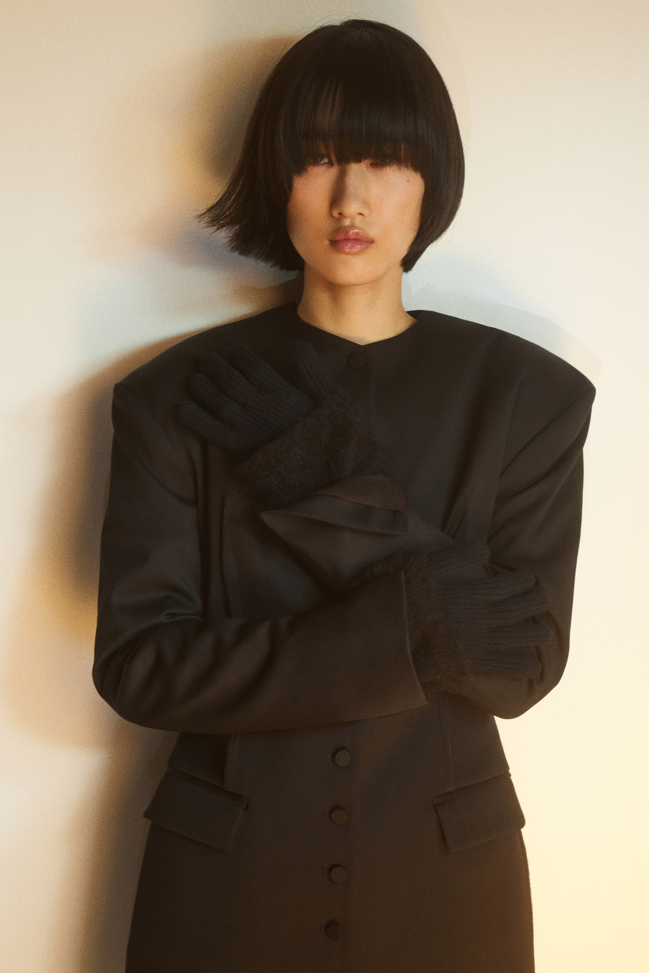 이미지 확대 보기: 부드럽고 섬세한 니트 소재에 질감 있는 립 커프스가 더해진 블랙 장갑을 모델이 착용하고 있으며, 구조적인 블랙 코트를 입은 채 팔을 가슴 앞에서 교차한 포즈를 취하고 있습니다. 장갑은 손목까지 올라오며 코트 소매 아래로 자연스럽게 레이어드되어 있습니다.