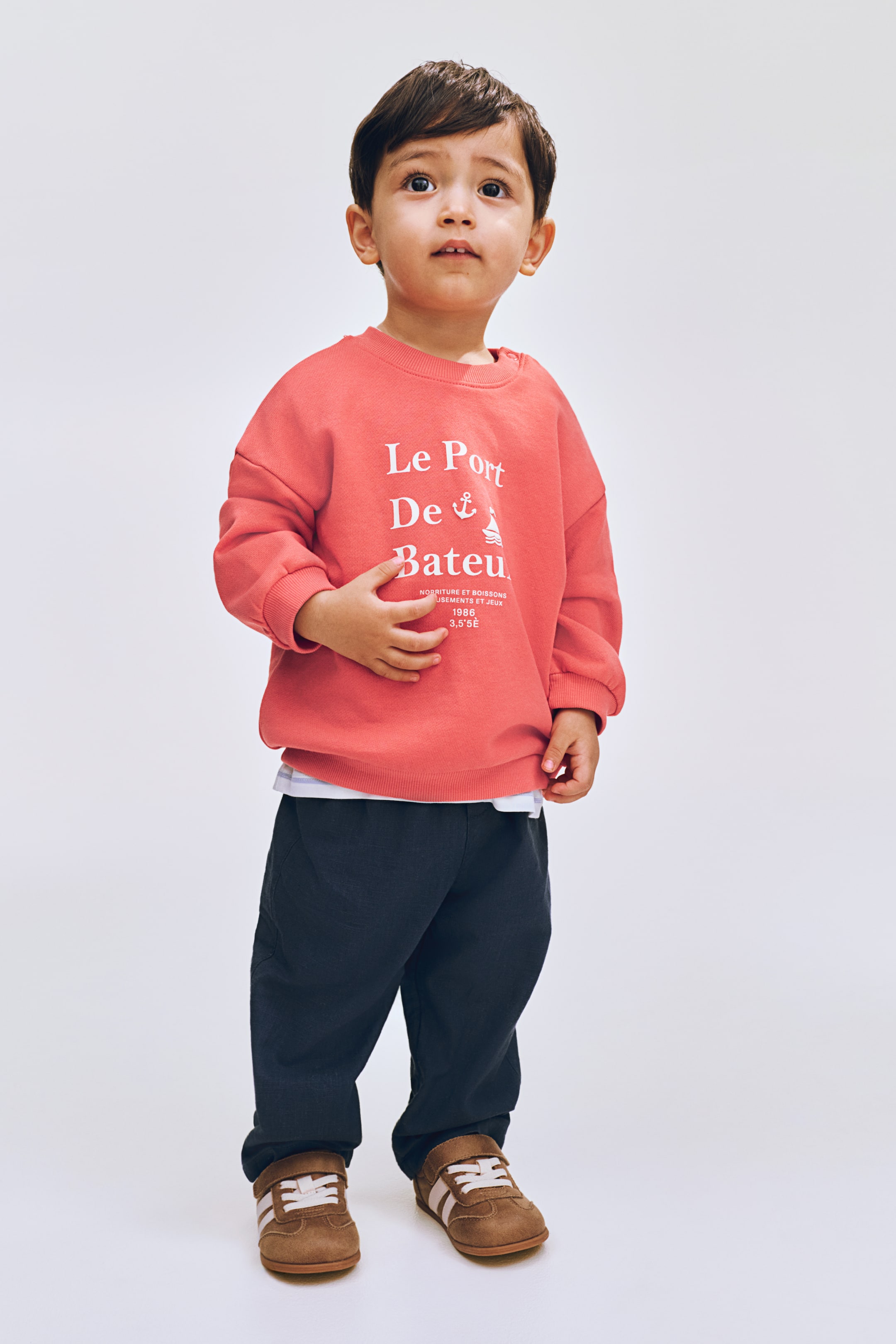 View larger image: A coral crewneck sweatshirt is showcased from the front, featuring long sleeves with ribbed cuffs and a ribbed hem, while a white t-shirt subtly emerges from beneath the hem. The front displays a white graphic print with the text "Le Port De Bateau," an anchor, and a boat symbol, accompanied by smaller text underneath.