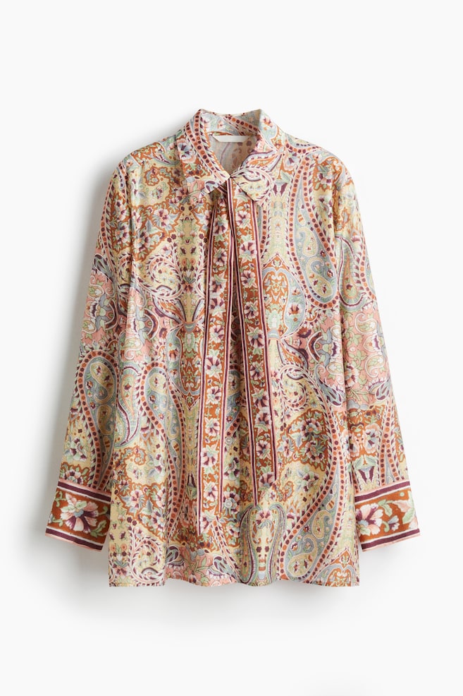 Ein langärmliges Hemd mit Vorderansicht zeigt ein lebhaftes Paisley- und Blumenmuster in Braun-, Orange-, Blau-, Grün- und Pinktönen. Es verfügt über einen spitzen Kragen und ein Krawattenhals-Detail mit zwei Stoffstreifen, die vorne herunterhängen, und seine langen Ärmel enden in breiten Manschetten mit einem kontrastierenden Randmuster. - 1