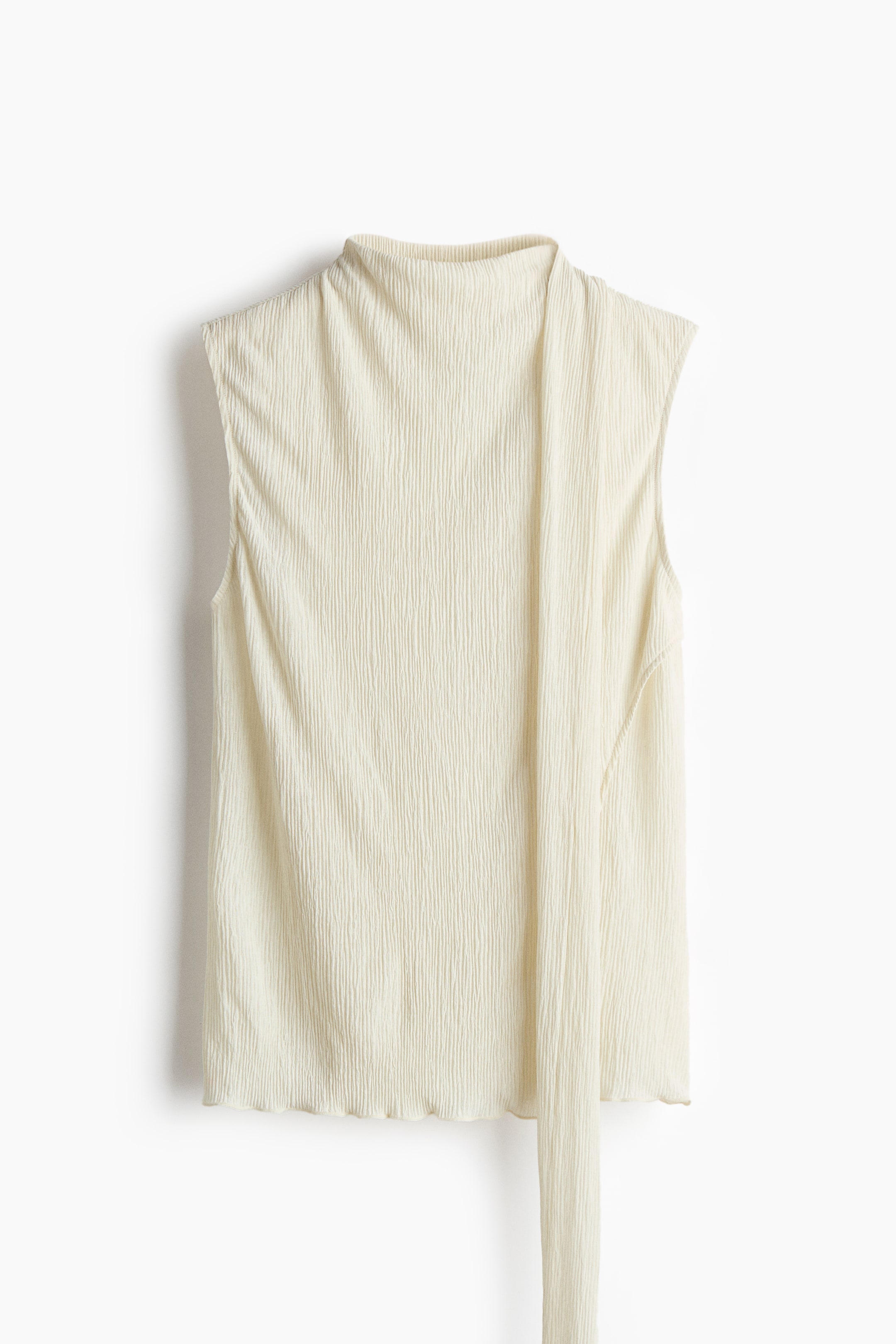 View larger image: The front of an off-white sleeveless top features a textured, crinkled fabric. It displays a mock-neck, a long tie detail extending from the right side, and a subtly ruffled hem.