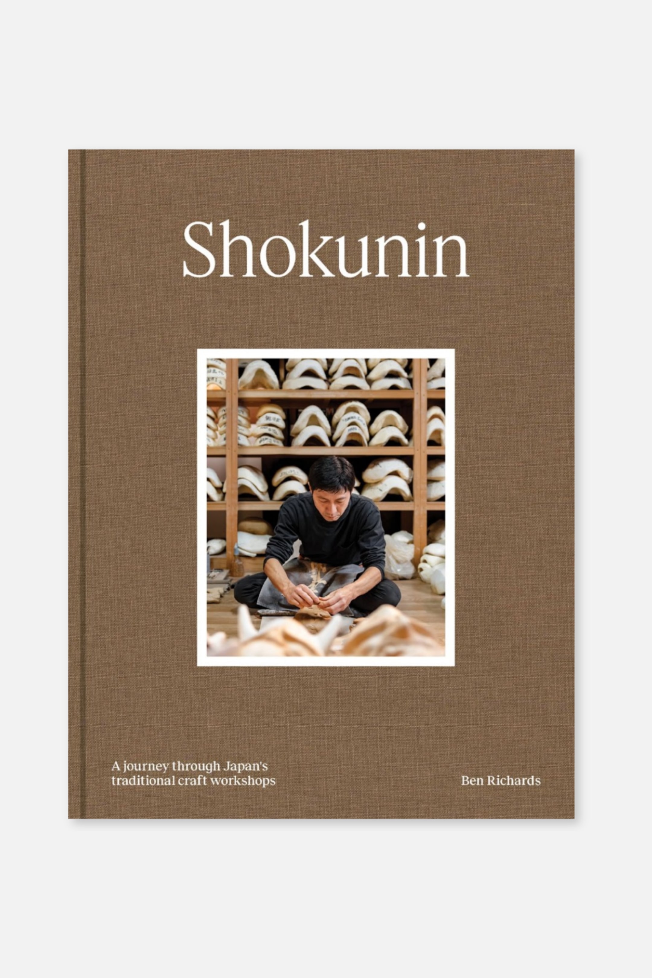 Agrandir l'image: Un livre présente sa couverture avant, qui arbore un matériau brun texturé avec le titre "Shokunin" imprimé en lettres majuscules blanches en haut. Une photographie rectangulaire montrant une personne travaillant sur des objets artisanaux est centrée sous le titre, et le sous-titre "Un voyage à travers les ateliers d'artisanat traditionnel du Japon" ainsi que l'auteur "Ben Richards" sont imprimés en texte blanc en bas.