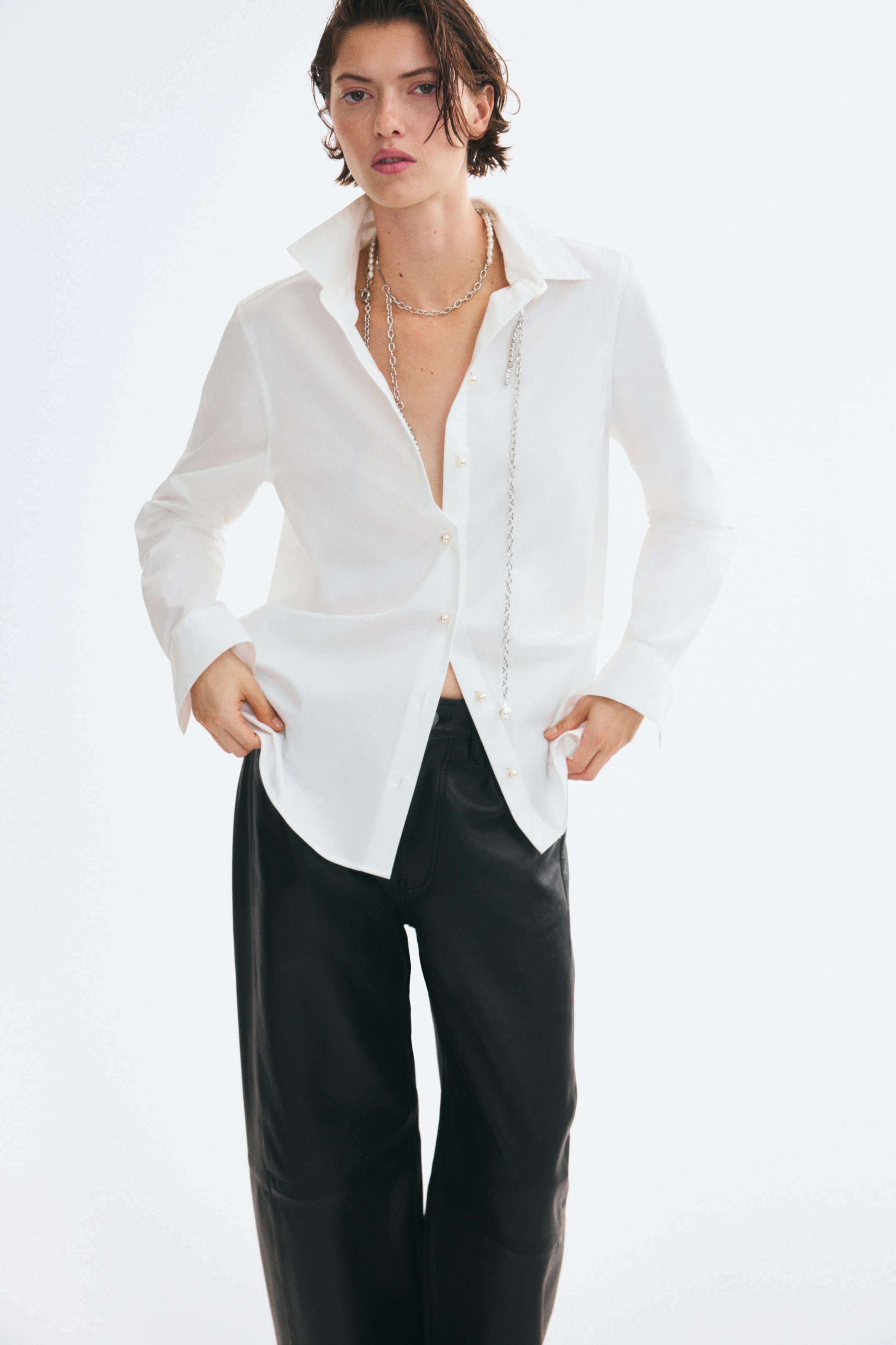 View larger image: A white cotton twill shirt is being worn by the model, who is holding the sides of its rounded hem. The shirt features a pointed collar, long sleeves, and pearly front buttons left partially undone, and is styled with black trousers and silver-toned necklaces.