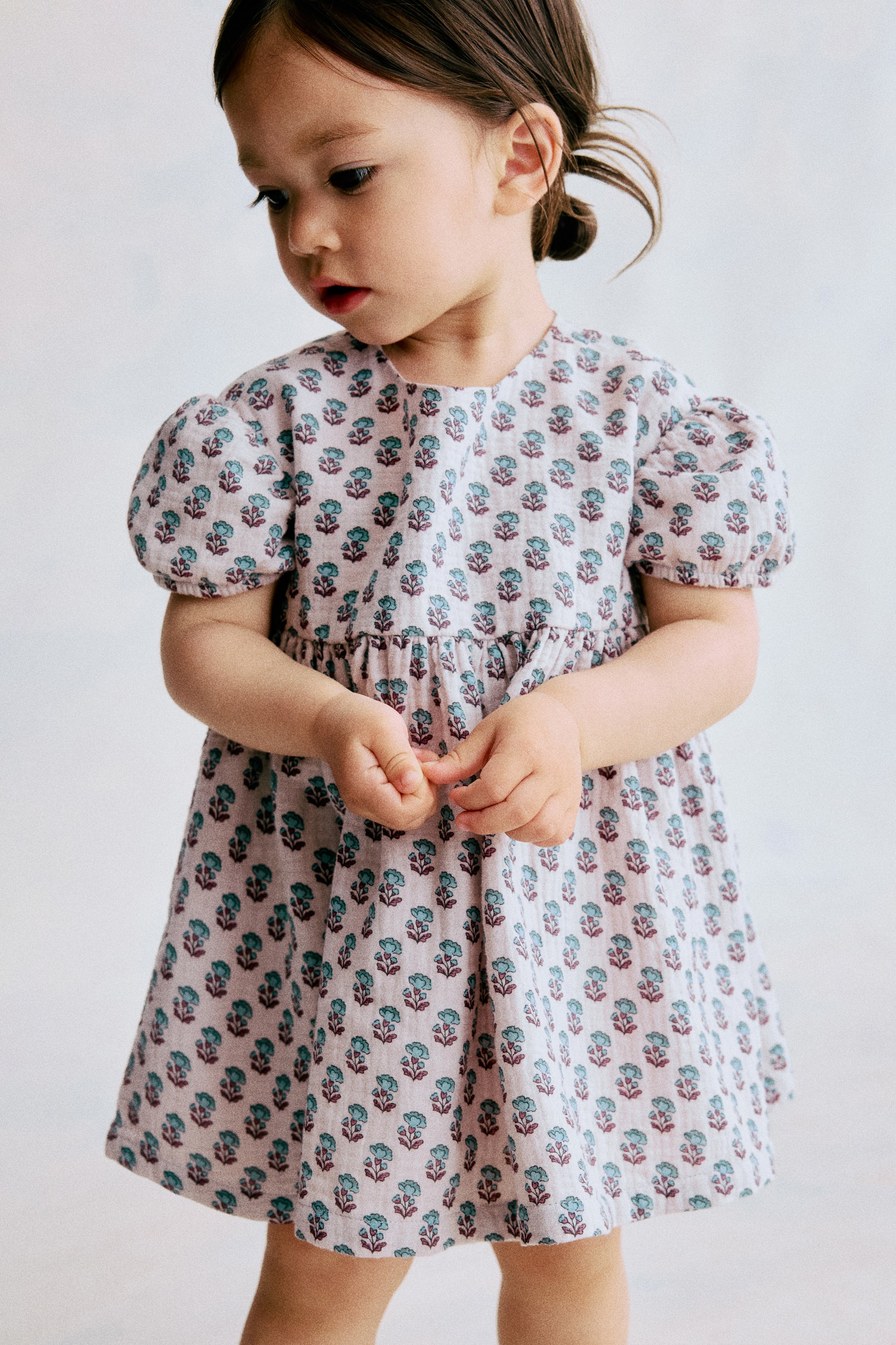 View larger image: A light-coloured dress is shown front-facing, featuring a repeating teal and maroon floral pattern. It is designed with a round neck, short puffed sleeves, and a gathered waistline that flows into a flared skirt.