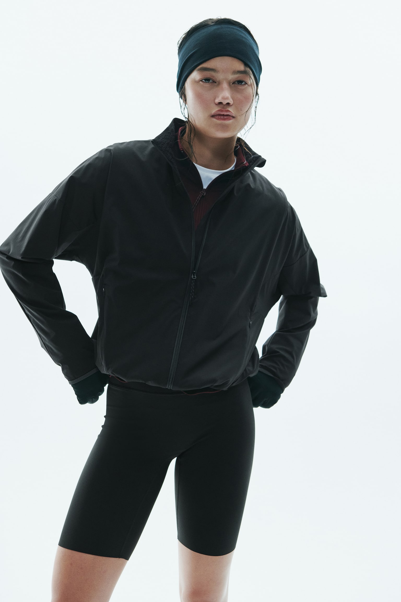 A black activewear jacket featuring a stand collar, a front zip fastening, and dropped shoulders is being worn by the model. The jacket is layered over a dark red top and styled with black athletic shorts, gloves, and a headband. - 1