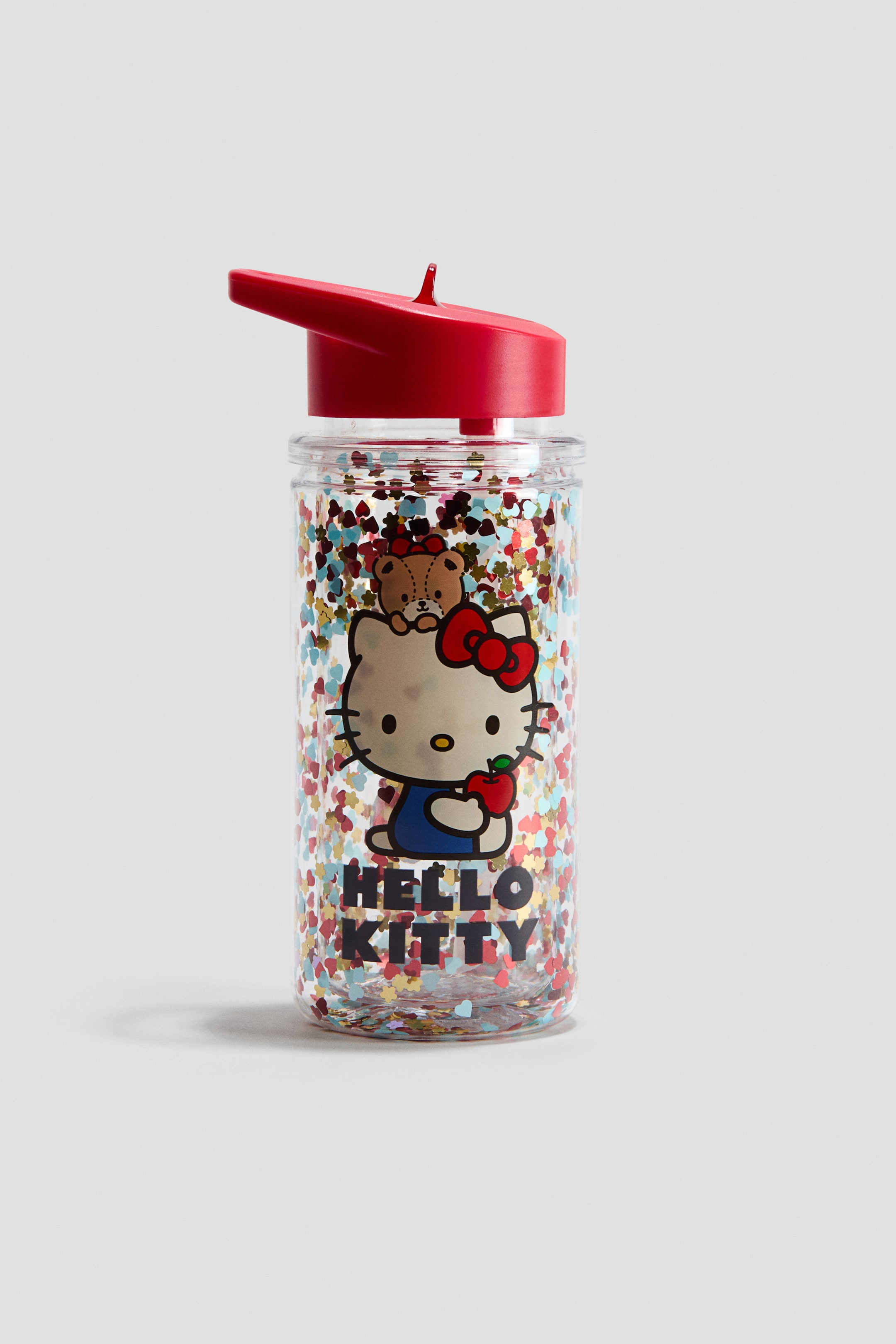 Ver imagen más grande: Una botella de agua de plástico transparente presenta un personaje de Hello Kitty de frente, vistiendo un vestido azul y un lazo rojo, sosteniendo una manzana roja, con un pequeño osito de peluche marrón apareciendo detrás de su cabeza, y el texto "HELLO KITTY" debajo. La botella contiene confeti en forma de corazones multicolores y está coronada con una tapa roja que incorpora una pajita y un pequeño asa.