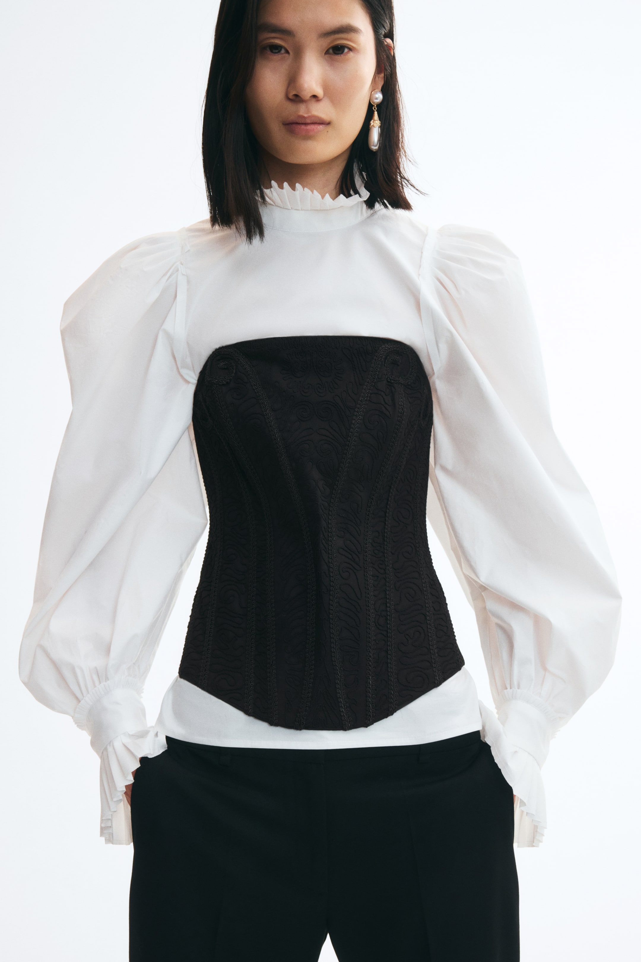Ver imagen más grande: La modelo lleva un top corsé bandeau negro con un intrincado bordado en tono y una estructura de varillas verticales. El top sin tirantes se superpone sobre una blusa blanca de cuello alto con voluminosas mangas abullonadas y se combina con pantalones negros.