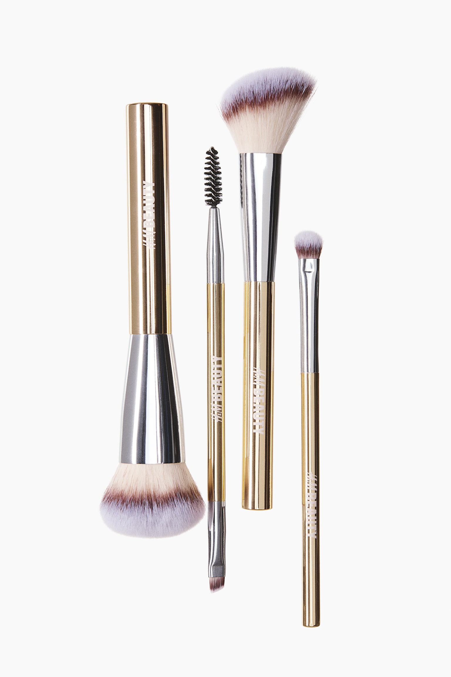 A set of four makeup brushes is presenting metallic gold-toned handles, silver ferrules, and synthetic bristles with a cream-to-purple gradient. The assortment is featuring a large powder brush, a dual-ended brow brush with a spoolie, an angled cheek brush, and a small eyeshadow brush.