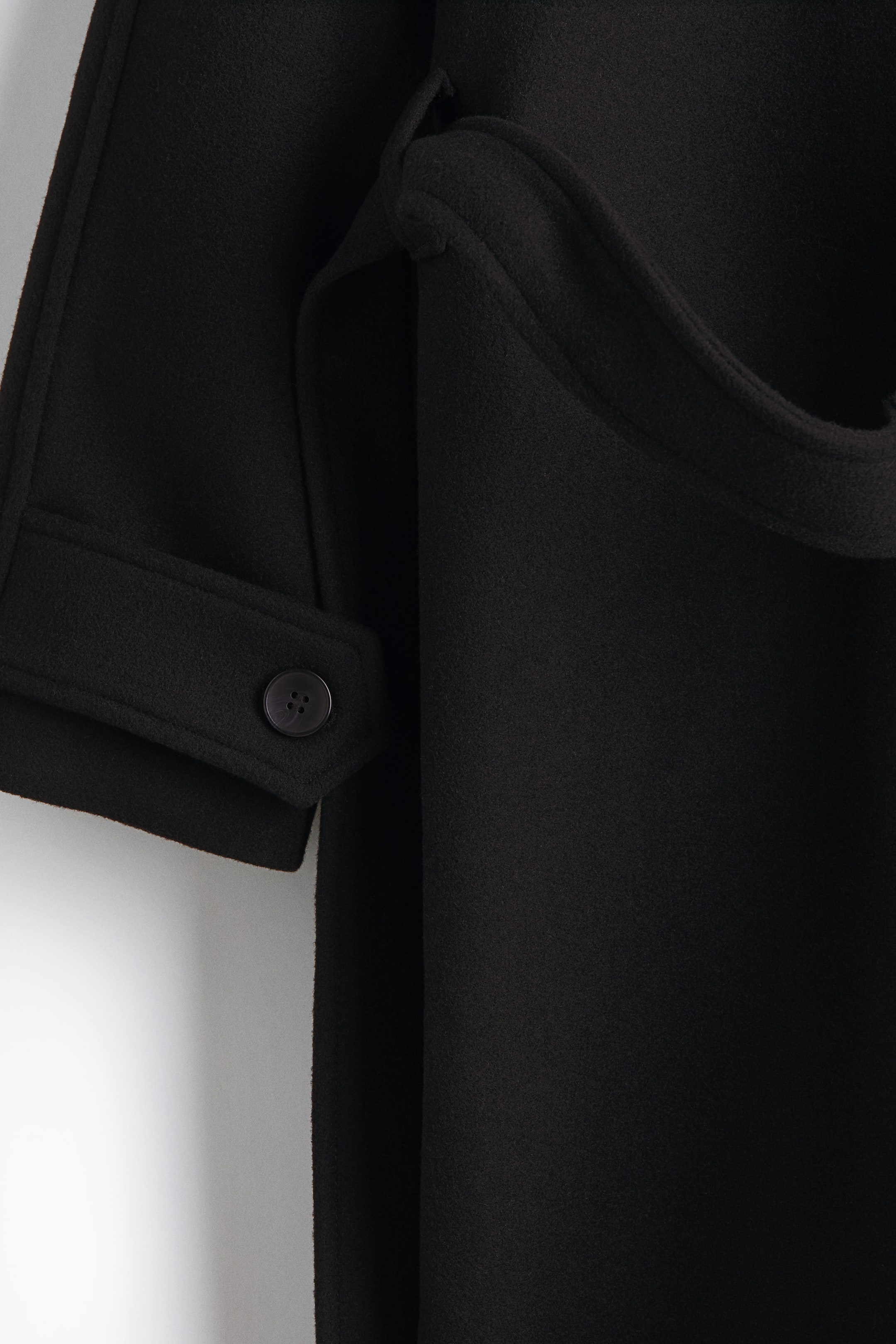 拡大画像を表示: 黒のコートは、ボタン付きのカフタブで固定された袖が特徴です。ベルトに似た広くて曲がった生地の帯が、衣服の右側に折りたたまれた状態で見えています。