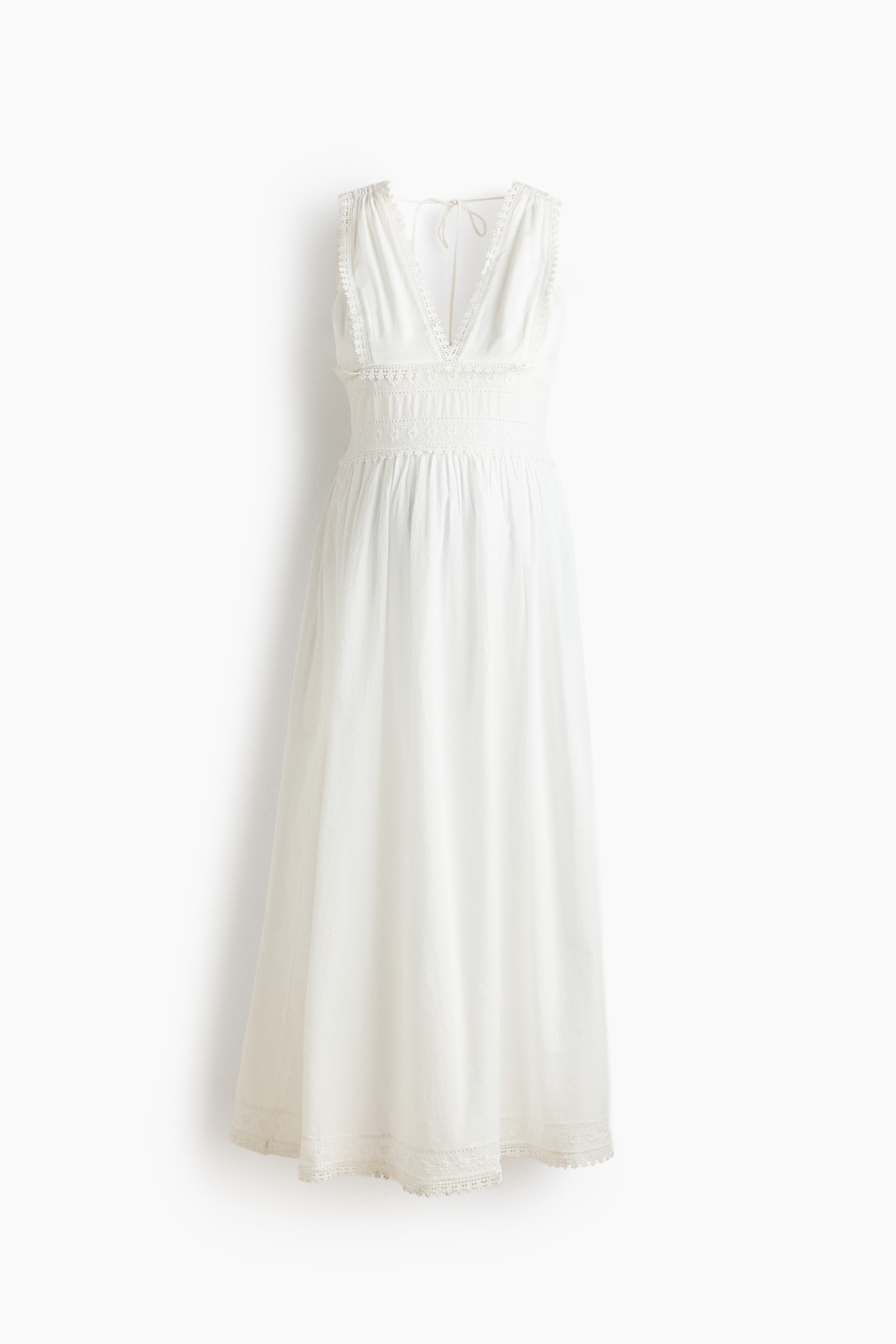 View larger image: A white maxi dress is showcased in its front view, featuring a deep V-neckline, sleeveless design, and delicate lace trim along the neckline, armholes, and hem. The bodice displays smocked detailing below the bust, transitioning into a flowing skirt, with ties visible at the back of the neck.