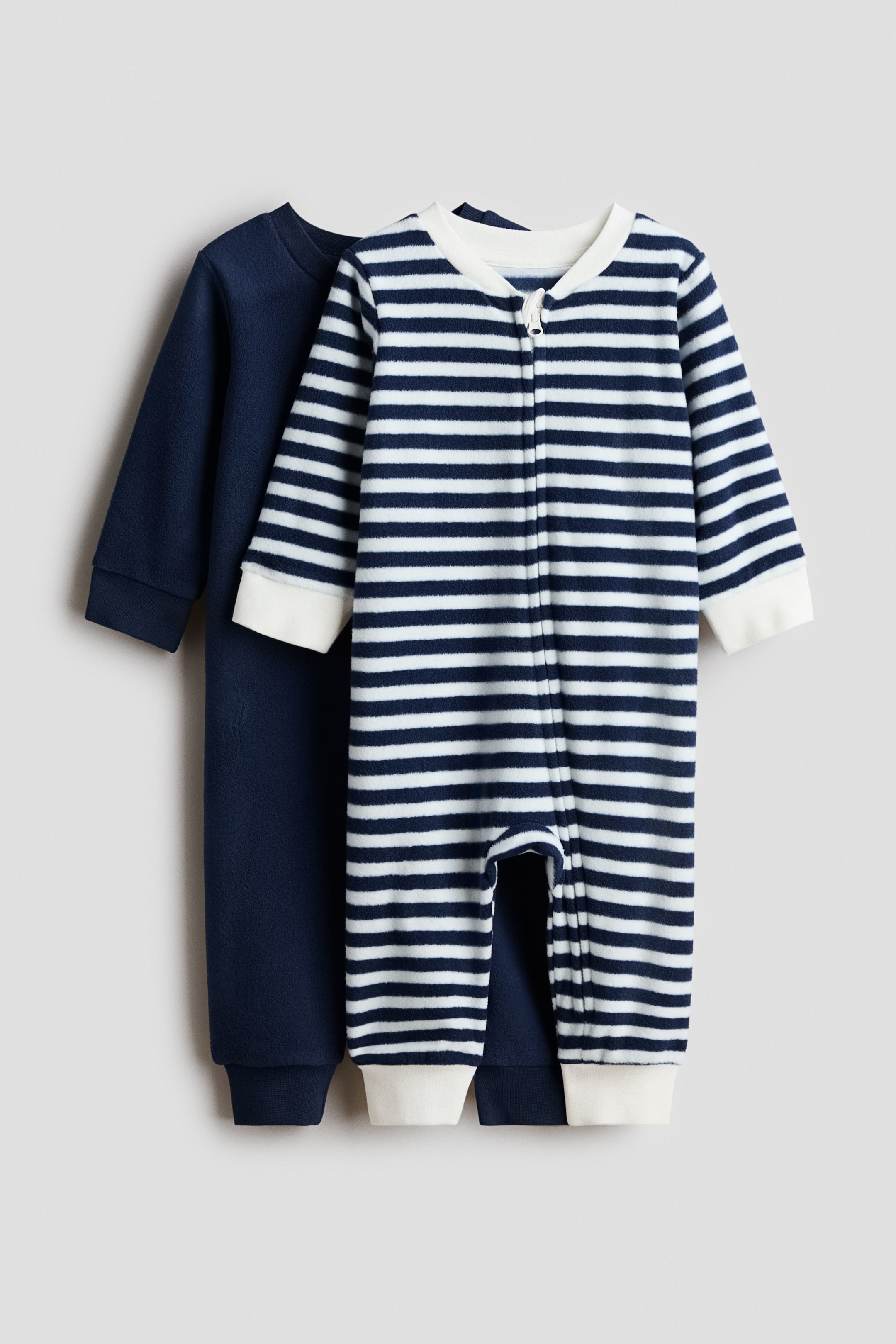 View larger image: Two baby fleece sleepsuits are showcased, with a solid navy blue sleepsuit positioned behind a striped one. The front sleepsuit features white and navy horizontal stripes, a full-length white zipper, and white ribbed cuffs at the wrists and ankles, while the navy sleepsuit displays matching navy cuffs.