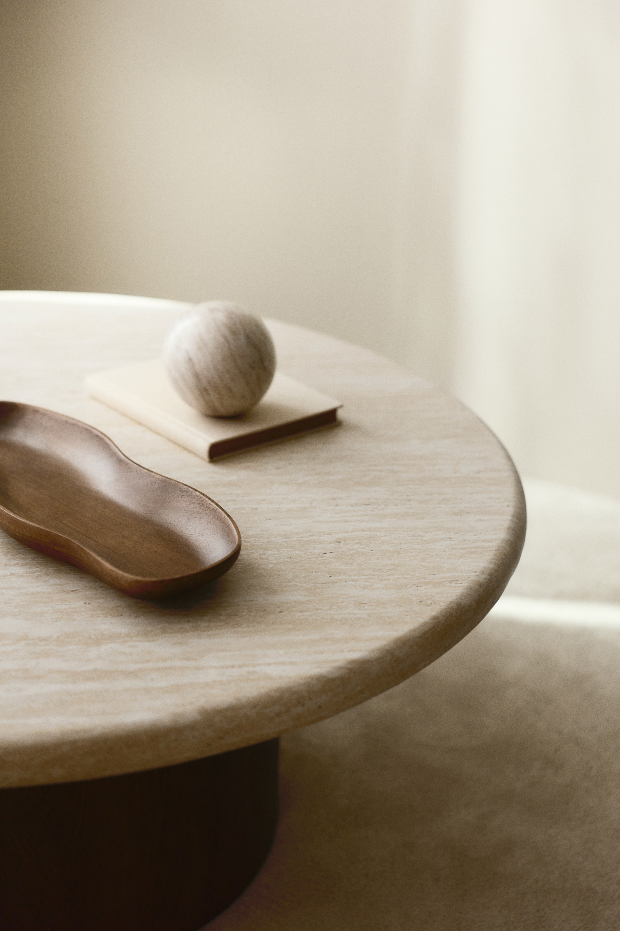 Agrandir l'image: La table ronde présente un plateau texturé de couleur claire, semblable au travertin, et un socle cylindrique foncé, vue d'un angle légèrement surélevé. Un plateau en bois foncé en forme de rein et une sphère de couleur claire reposant sur un petit livre de couleur claire sont positionnés sur la surface de la table.
