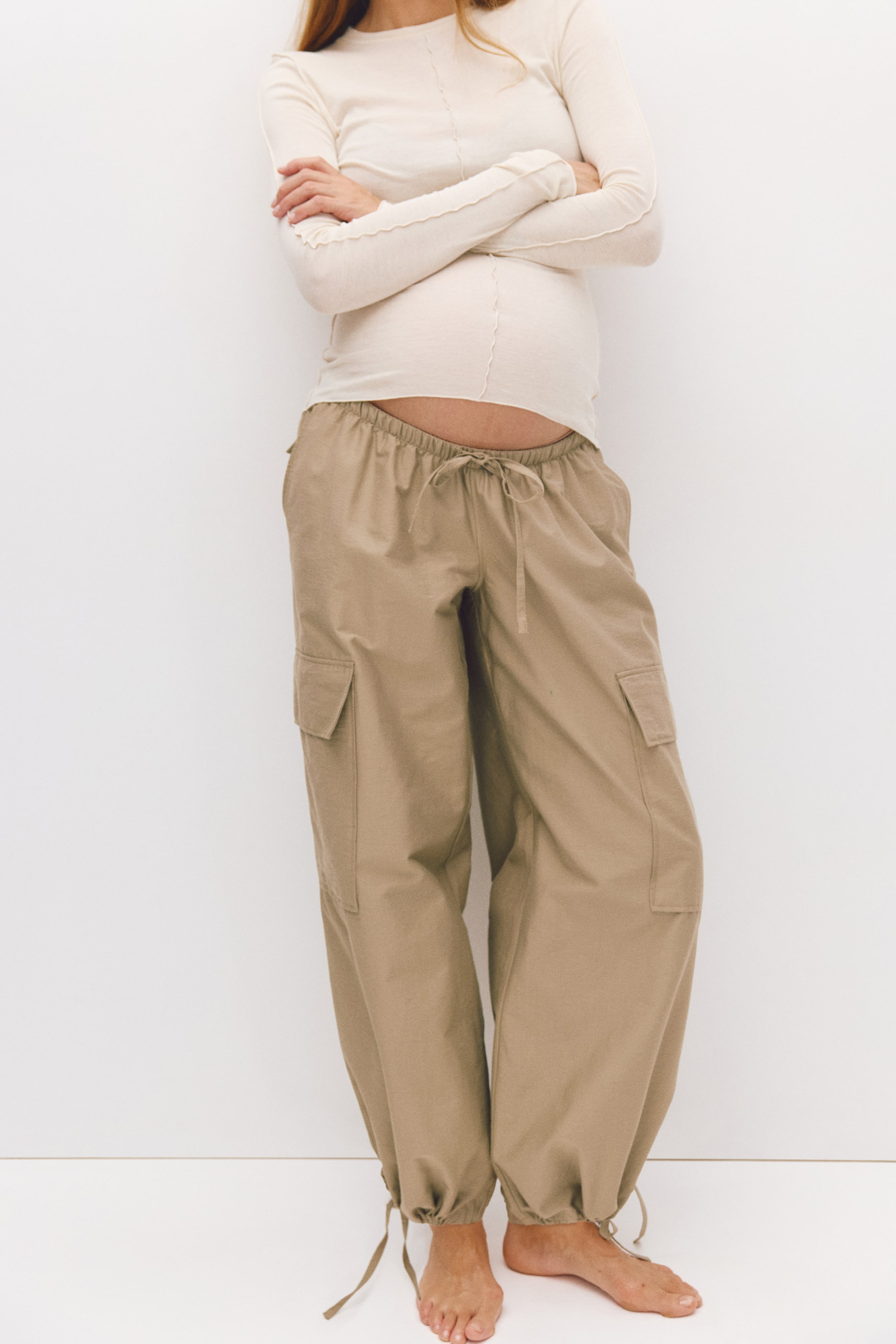 View larger image: The model is wearing relaxed-fit cargo trousers in a tan cotton blend, styled with a long-sleeve cream top. The trousers feature a low, elasticated waistband with a tied drawstring, side cargo pockets, and balloon legs with adjustable drawstrings at the ankles.
