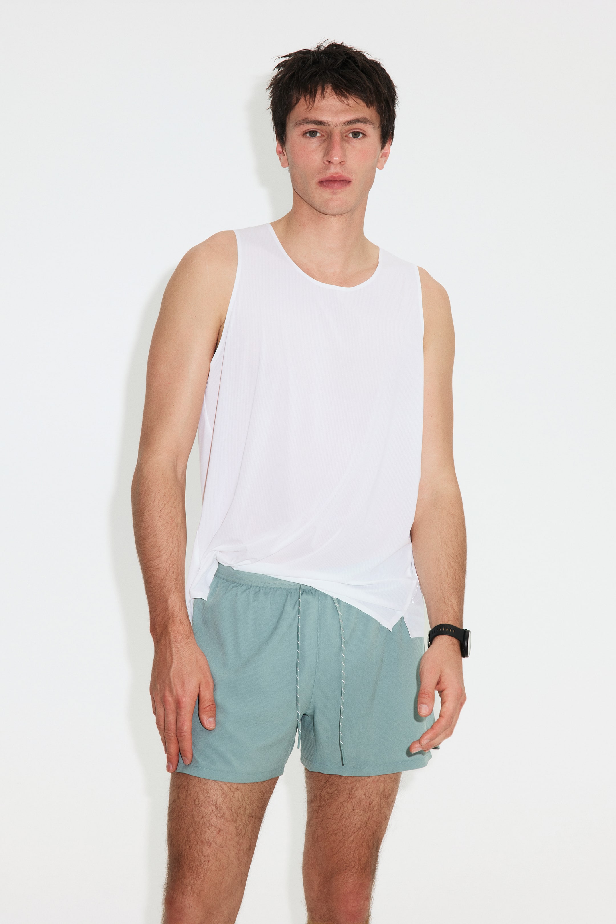 Visualizza immagine più grande: Una canotta bianca senza maniche, leggera e con una texture sottile, scende morbidamente su pantaloncini sportivi color teal chiaro. I pantaloncini presentano una fascia elastica in vita e un laccio bianco annodato sul davanti.