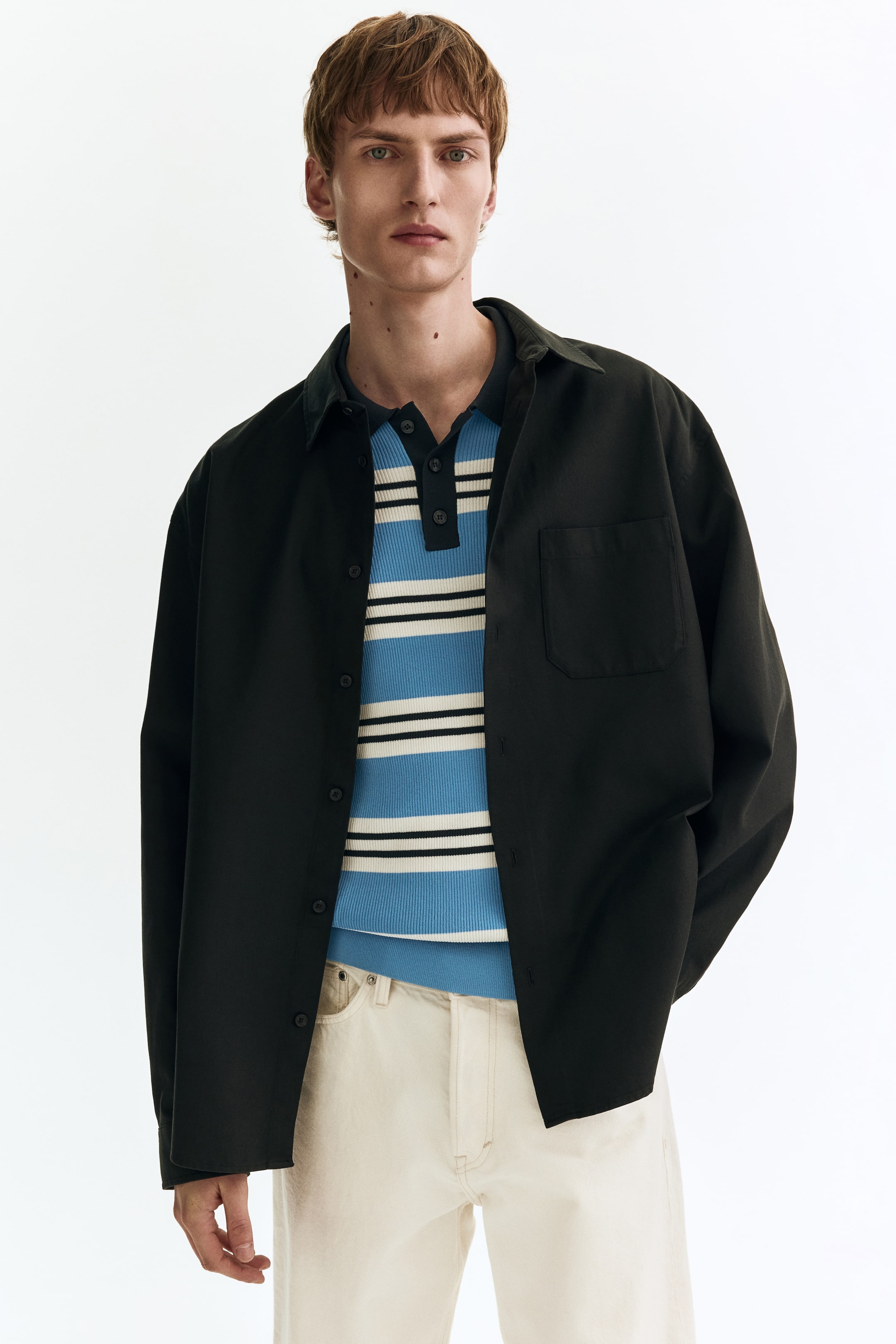 הצגת תמונה גדולה יותר: חולצה שחורה, בגזרה רפויה, עם שרוולים ארוכים וכפתורים, נלבשת פתוחה מעל חולצת פולו מפוספסת. היא מציגה צווארון פתוח, שורת כפתורים מלאה עם כפתורים כהים, וכיס טלאי יחיד על החזה השמאלי.