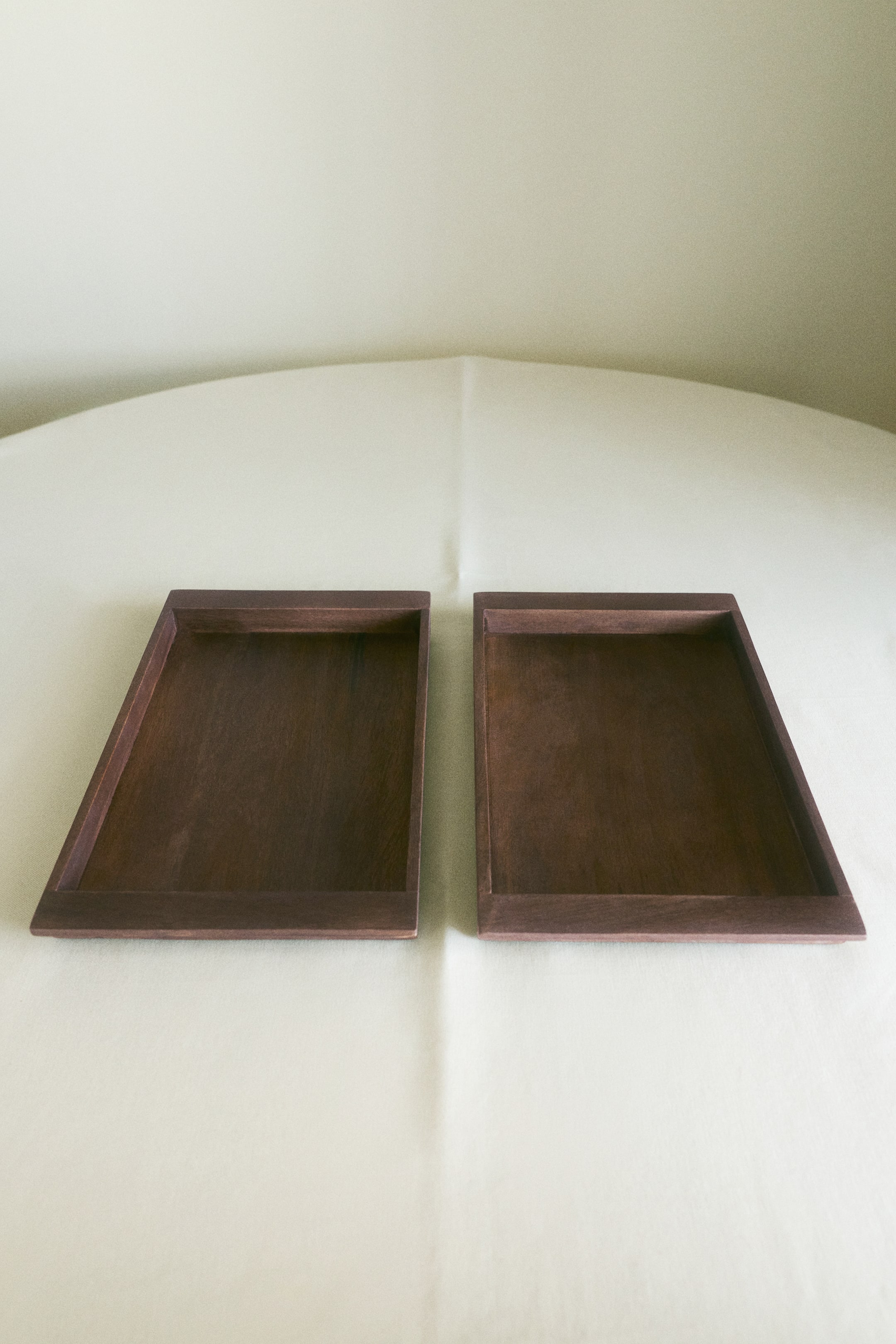 View larger image: Two rectangular serving trays in dark brown mango wood are lying side by side, viewed from the front. Each tray features a flat interior surface, a raised outer rim, and a visible natural wood grain texture.