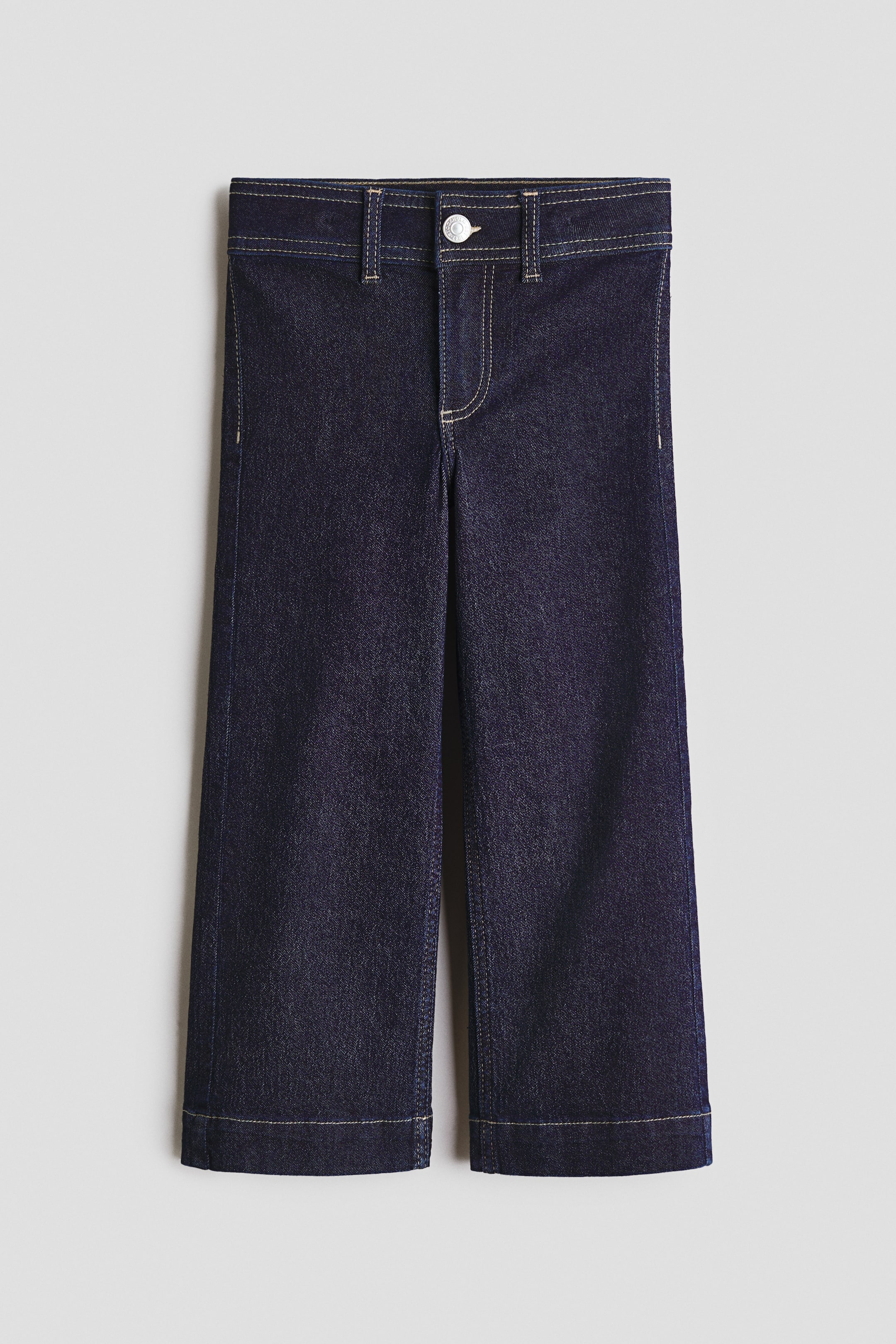 Ver imagen más grande: Pantalones vaqueros azul oscuro que presentan una vista frontal con una cintura plana, trabillas para el cinturón y un único cierre de botón plateado sobre una bragueta con cremallera. Las costuras de color claro en contraste delinean todas las costuras, incluyendo las perneras anchas y el dobladillo con vuelta.