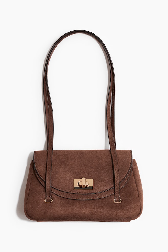 Un sac à main marron effet daim présente son devant, avec un rabat incurvé muni d'une fermeture à tourniquet dorée. Une longue et fine bandoulière est fixée aux côtés du sac par de petits anneaux dorés. - 1