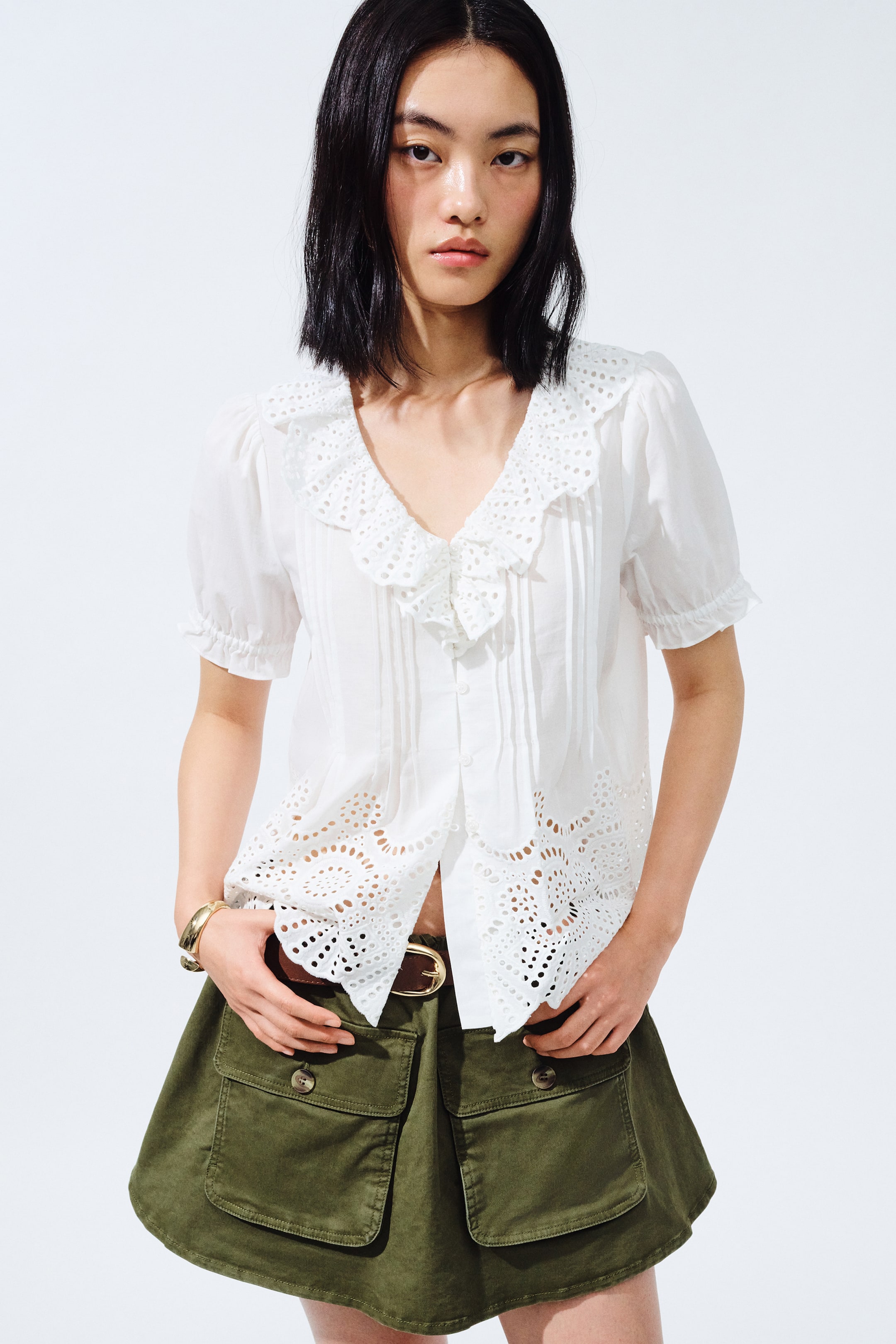 View larger image: The white blouse features a large, ruffled eyelet lace collar and short puffed sleeves with ruffled cuffs. Its front showcases vertical pintucks, and the scalloped hemline is designed with circular eyelet lace patterns.
