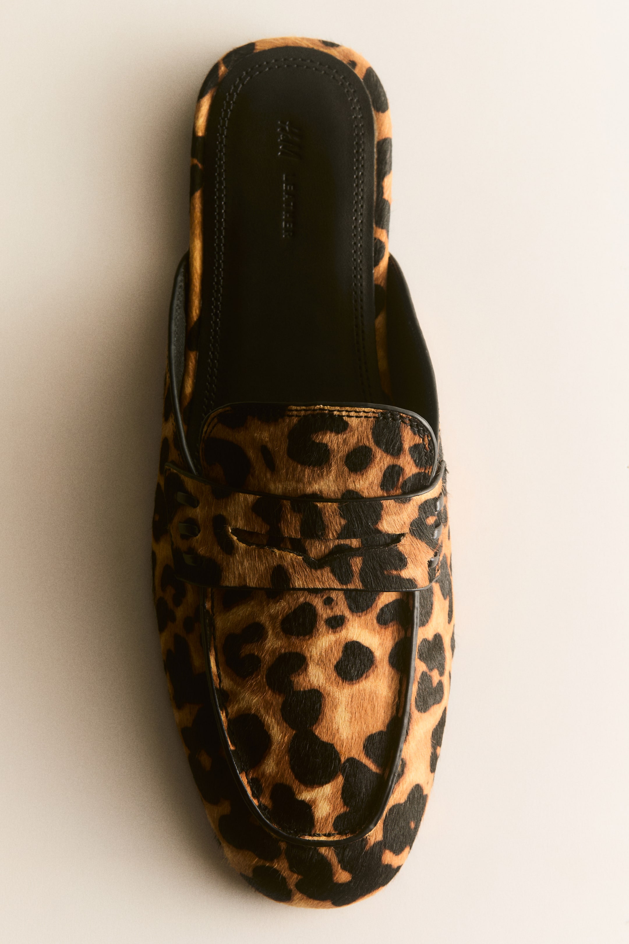 Ampliar la imagen: Un mocasín mule muestra un estampado de leopardo en su parte superior de cuero texturizado y afelpado. Presenta una punta almendrada, un adorno tipo "penny keeper" en la parte superior, una costura de mocasín en el frente y una plantilla visible de cuero negro.
