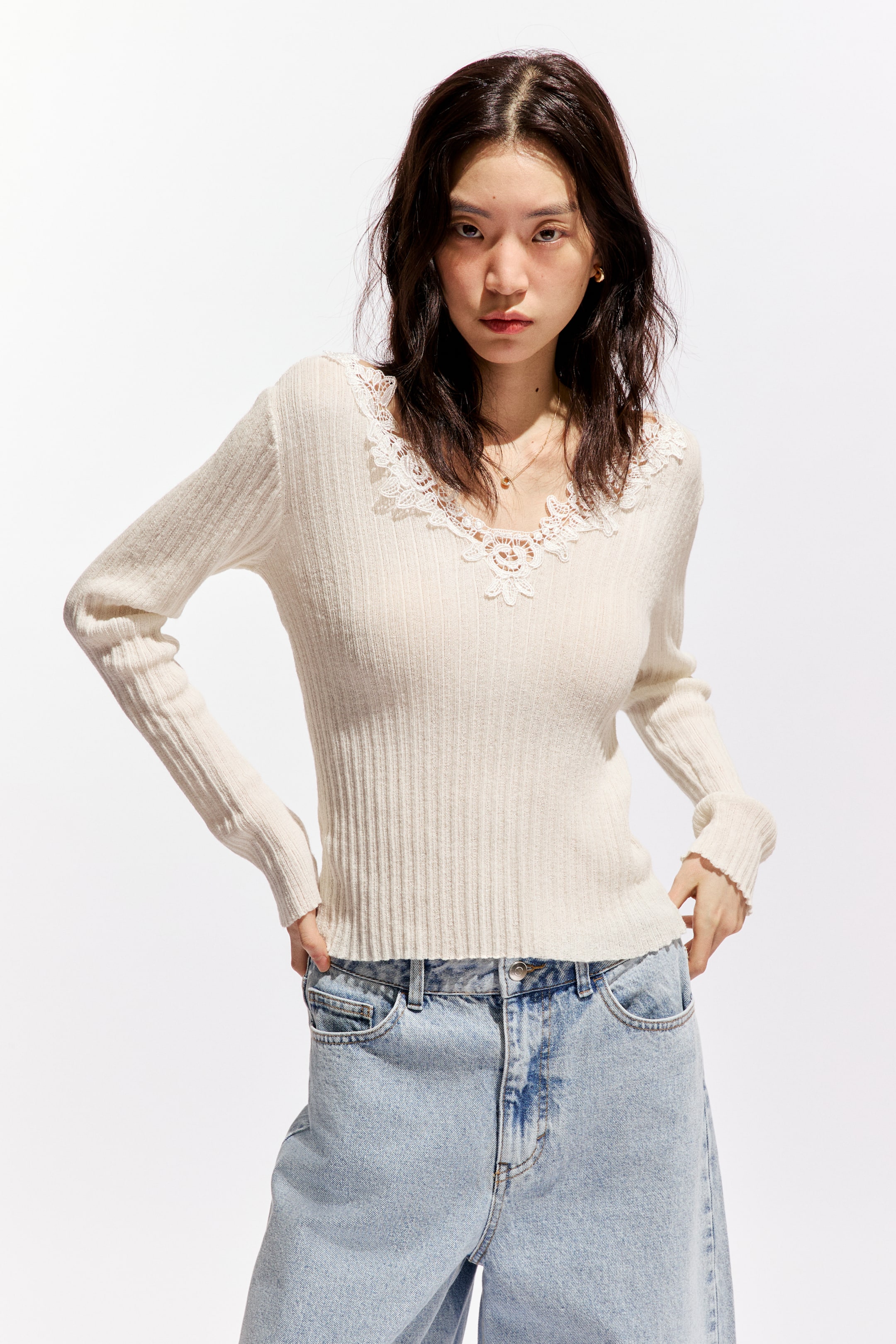 拡大画像を表示: クリーム色のリブニットトップが前向きに展示されています。Vネックラインには白い花柄のレーストリムが施され、長袖にはフリル付きのカフスが特徴です。