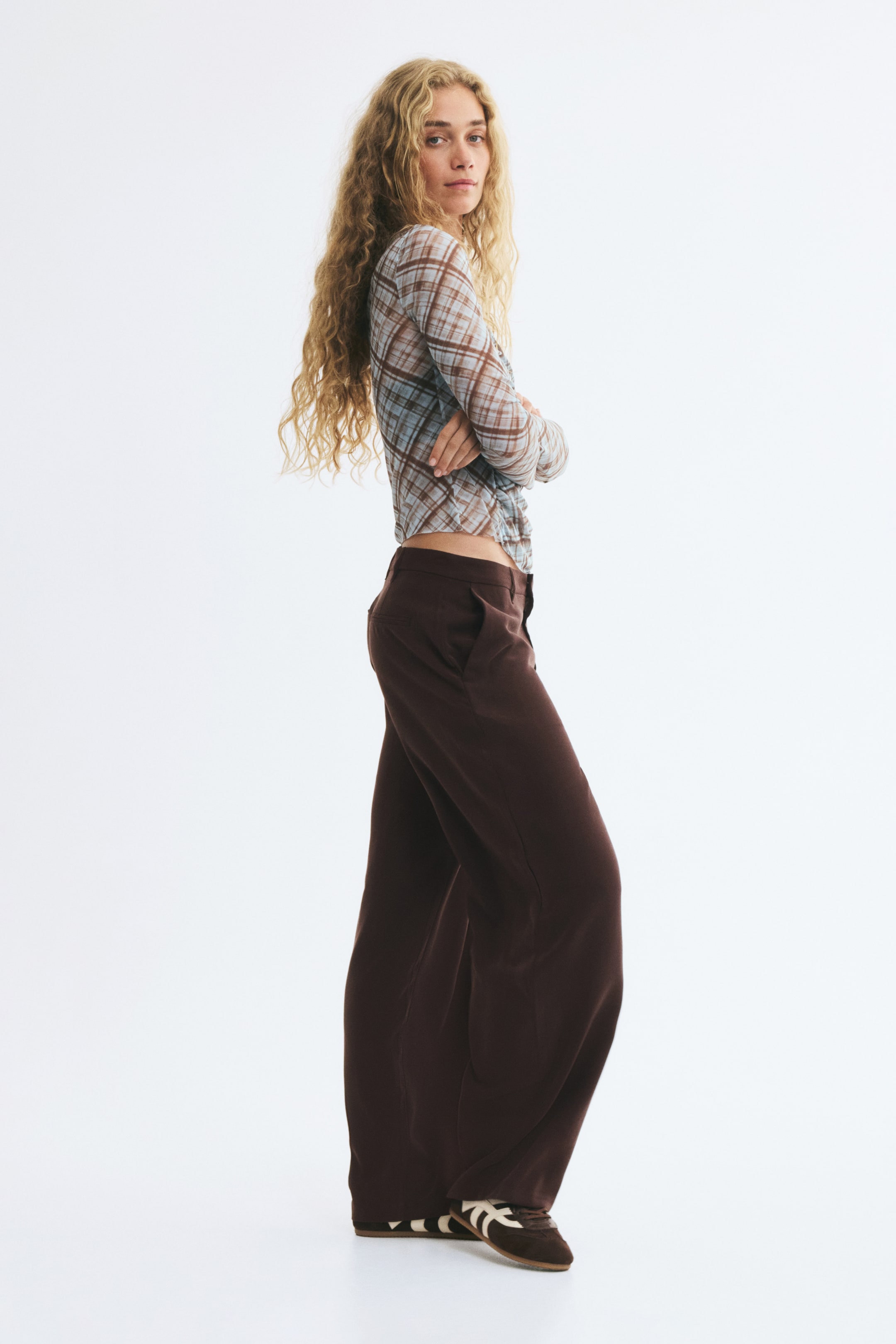 Prikaži veću sliku: Model nosi krojene tamno braon pantalone sa regularnim strukom, dijagonalnim prednjim džepovima i širokom barrel-leg siluetom. Pantalone su stilizovane uz providan, karirani top dugih rukava i niske patike.