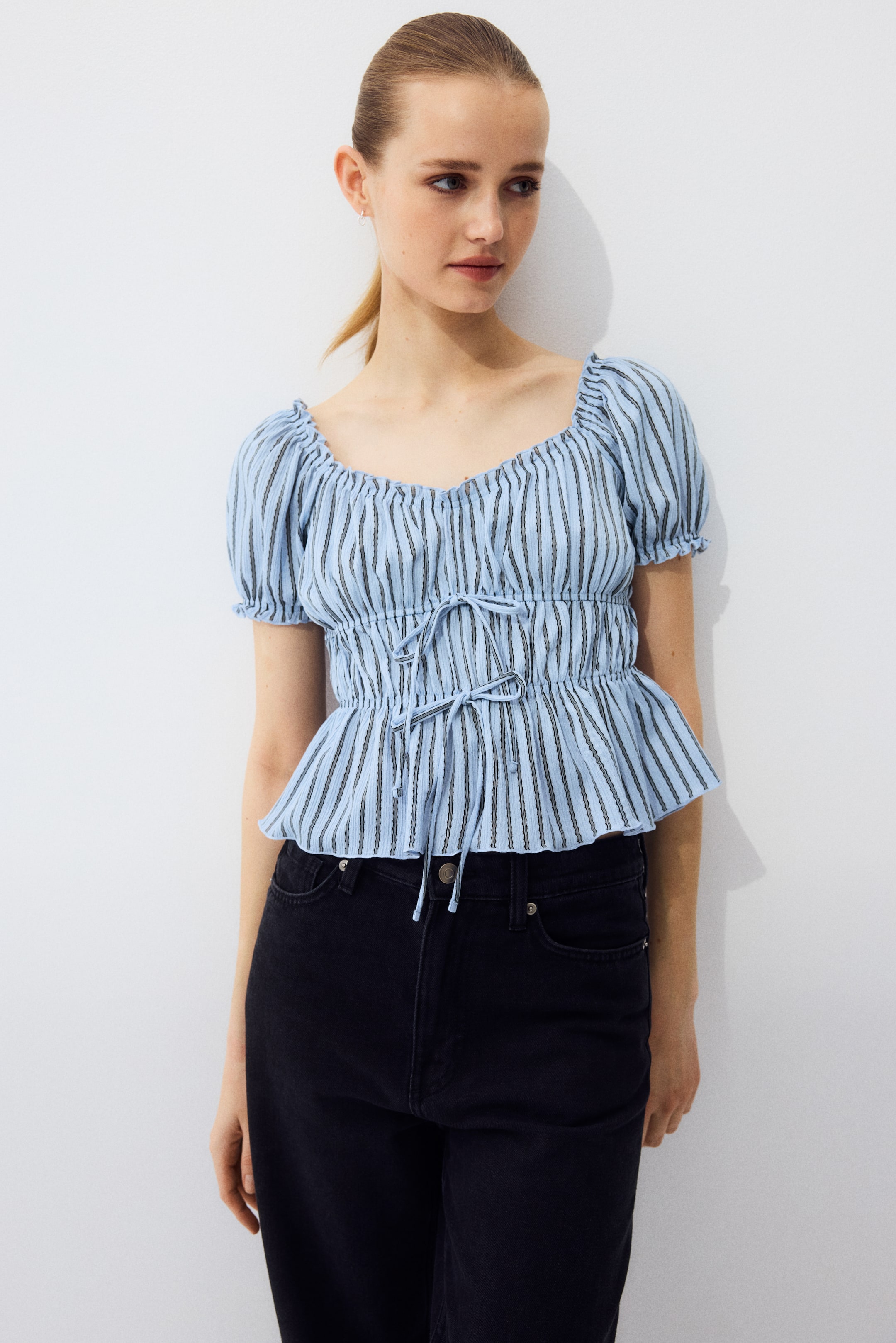 View larger image: The model is wearing a light blue textured, striped jersey top, featuring a deep, gently V-shaped neckline with a gathered frill trim. It has short puff sleeves with frill-trimmed cuffs, two elasticated seams with narrow ties at the waist, and a gently flared hem, styled with black jeans.