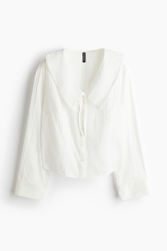 A white long-sleeved shirt, viewed from the front, is featuring a large, rounded sailor collar with a subtle textured trim along its edges. It is designed with a full button-front closure, a V-neckline with tie details, and long sleeves ending in simple cuffs, while its fabric is displaying a subtle vertical stripe texture. - 1