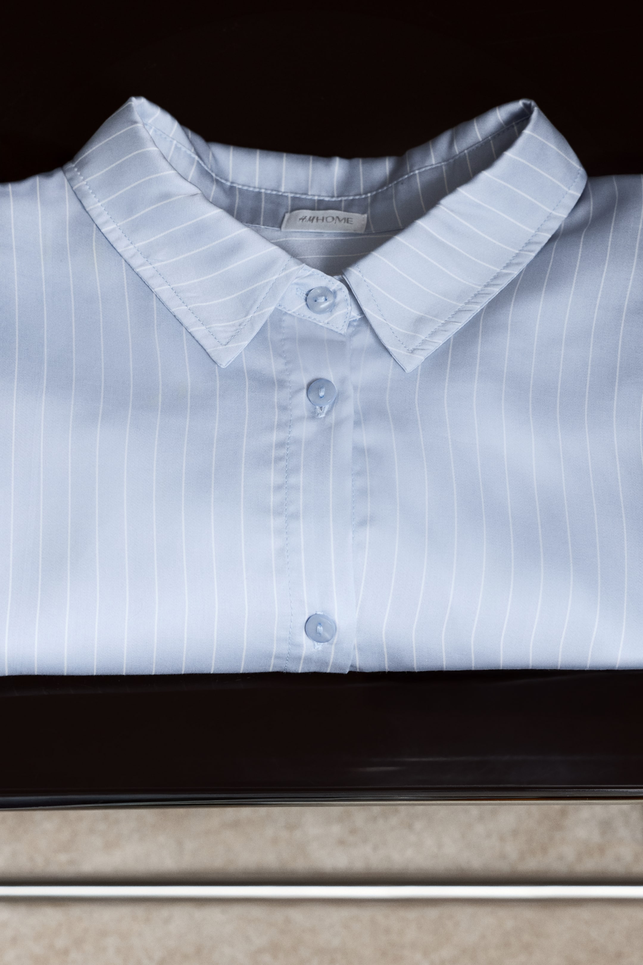 View larger image: A light blue shirt with white pinstripes is lying neatly folded, showcasing its pointed collar and three light blue buttons on the front placket. The "H&M HOME" tag is visible on the inner collar.