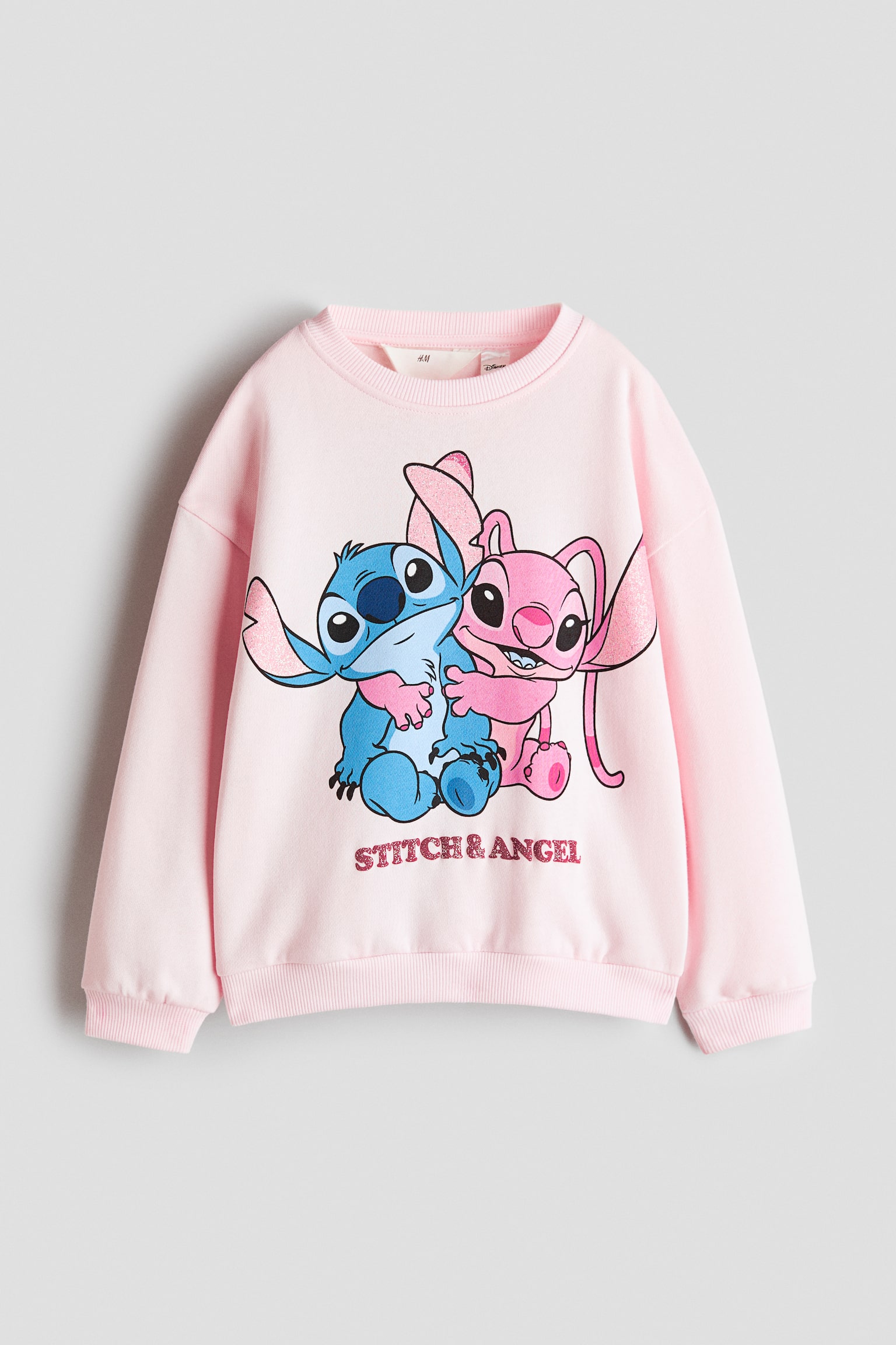 Un maglione oversize rosa chiaro Ã¨ mostrato frontalmente, con scollo rotondo rifinito a coste, spalle scese e bordo a coste su polsini e fondo. Sul davanti Ã¨ presente una grafica stampata con i personaggi dei cartoni animati Stitch e Angel che si abbracciano, con la scritta "STITCH & ANGEL" in caratteri rosa glitterati sotto di loro.