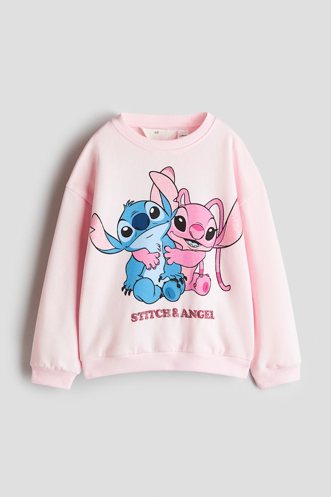 Un jersey oversize rosa claro aparece de frente, con cuello redondo acabado en canalé, hombros caÃdos y ribete de canalé en los puños y el bajo. En la parte delantera muestra un gráfico impreso de los personajes de dibujos animados Stitch y Angel abrazándose, con el texto "STITCH & ANGEL" en letras rosas con efecto glitter debajo. - 1