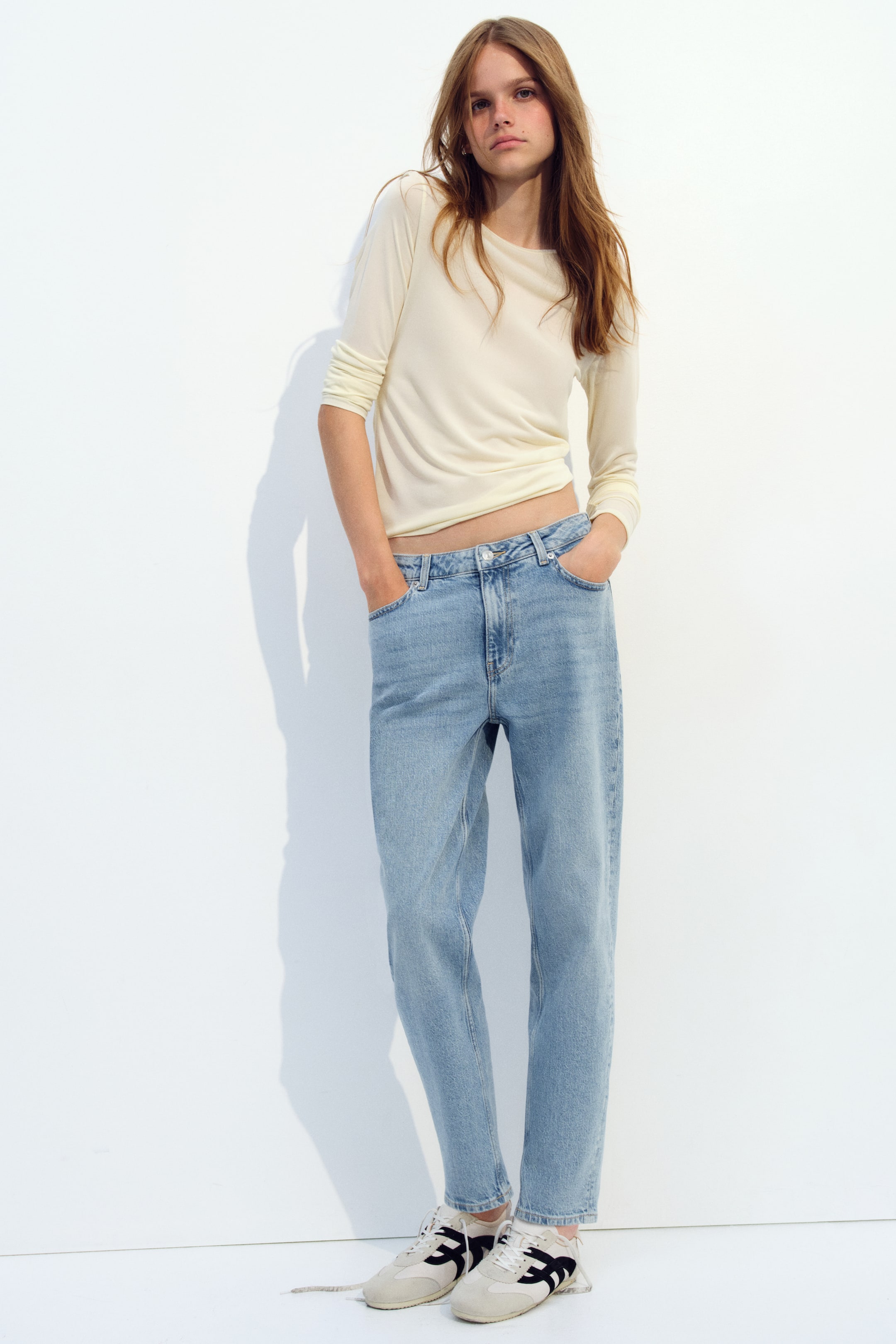 View larger image: Light-wash blue denim jeans are worn by the model, who is standing with hands in the front pockets. The high-waisted, five-pocket jeans feature a slim fit through the hips and thighs, tapering slightly at the ankle, and are styled with a cream long-sleeve top and low-top sneakers.