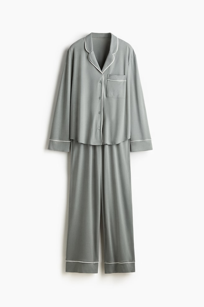 A set of grey pyjamas is hanging, presented FRONT. The long-sleeved shirt features white-piped notch lapels, a front button closure, and a chest patch pocket with white piping. The matching bottoms have wide, straight legs with white piping at the cuffs. - 1
