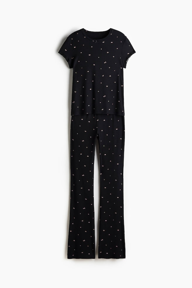 Musta pyjamasetti esitetään edestäpäin, ja siinä on ribattu modaalijersey-paita ja siihen sopivat housut. Paita on suunniteltu pyöreällä pääntiellä ja lyhyillä hihoilla, jotka on viimeistelty pitsireunuksilla. Housuissa on joustava vyötärö ja kevyesti levenevät lahkeet, ja molemmissa osissa on pieniä, hajanaisia vaaleanvärisiä kuvioita. - 1