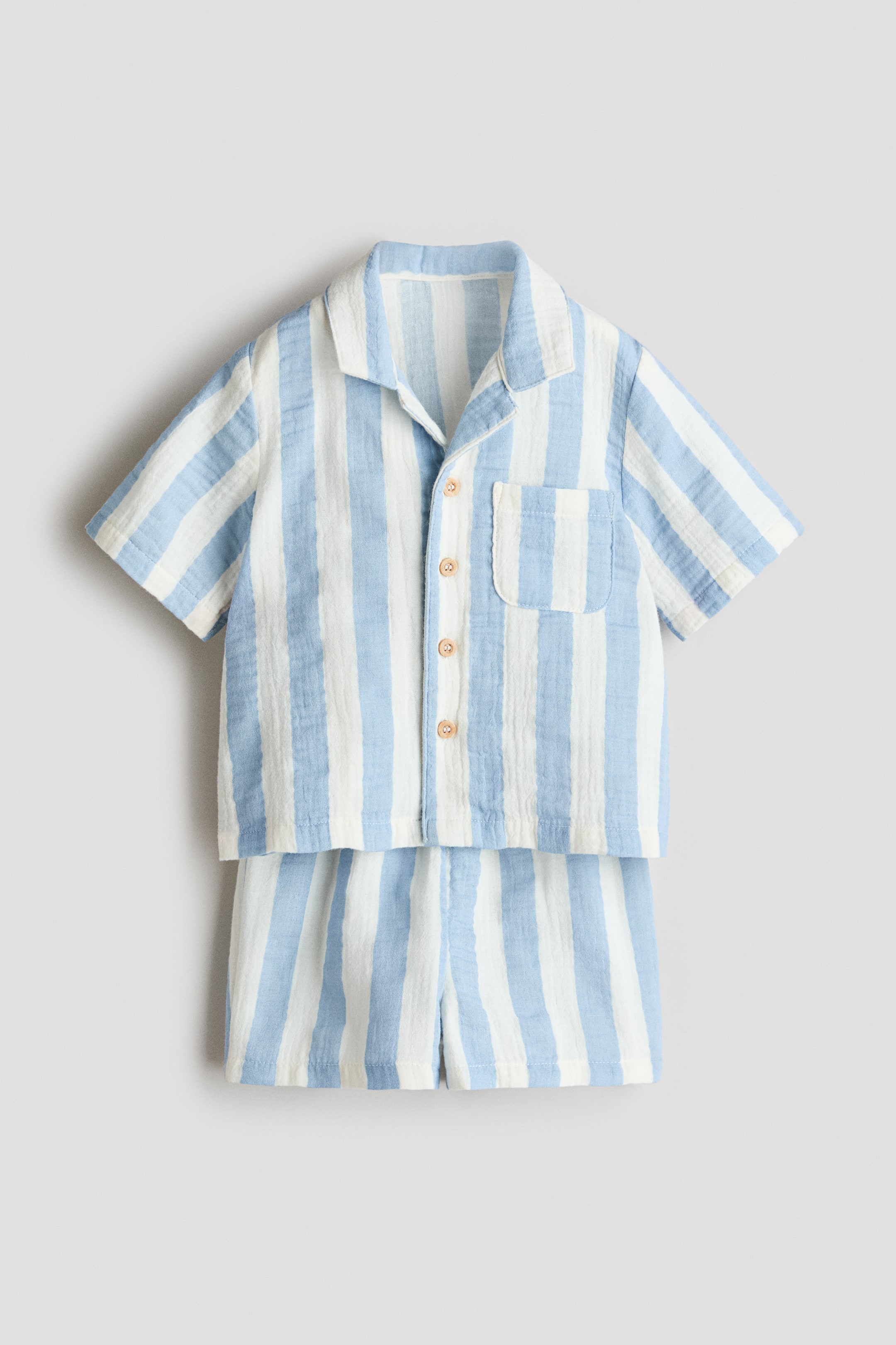 View larger image: A set of a shirt and shorts, displayed from the front, showcases vertical light blue and white stripes on a textured woven fabric. The straight-cut shirt features a resort collar, buttons running down the front, short sleeves, and an open chest pocket, while the shorts present a matching striped pattern.
