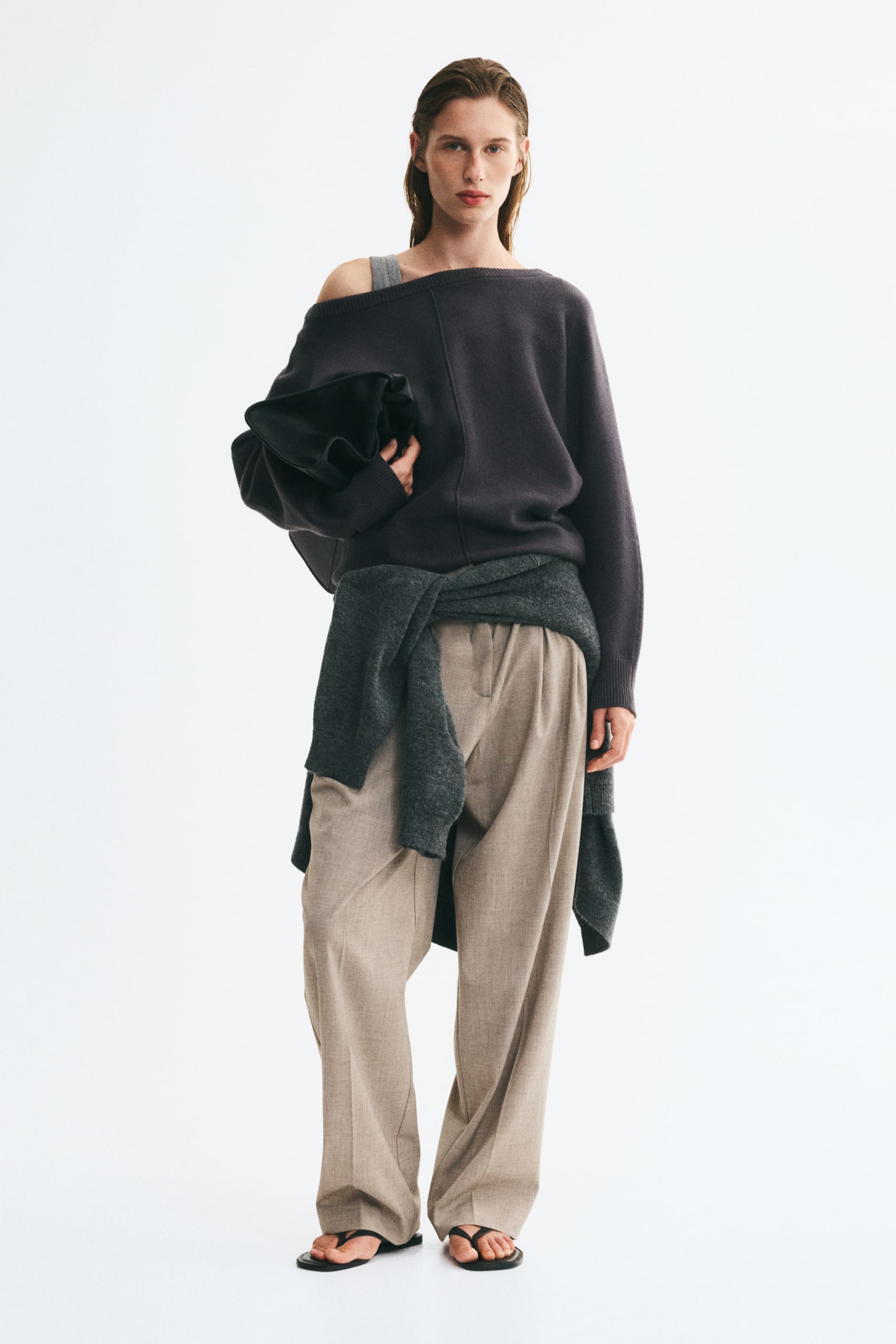 A dark grey knitted sweater featuring a rib-trimmed boat neck and a visible front seam is worn by the model, styled slightly off one shoulder. The long-sleeved sweater is paired with beige trousers, a second grey knit tied around the waist, and a black clutch bag held under the arm. - 1