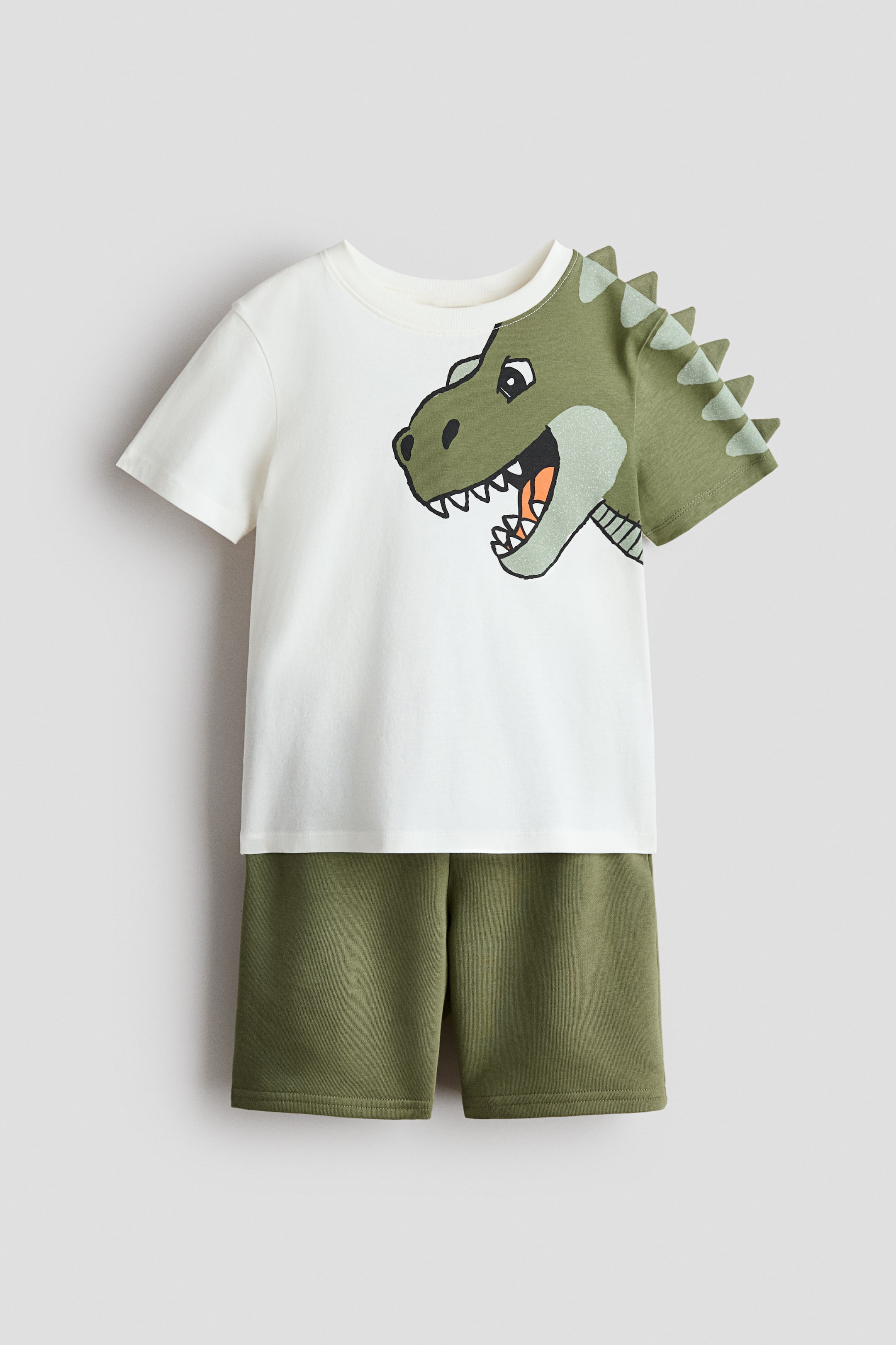 View larger image: A two-piece children’s set is shown from the front, consisting of a cream T-shirt and coordinating olive-green shorts. The T-shirt features a large green dinosaur head graphic on the front, with the graphic extending onto the right sleeve, which is also green and has raised spikes along the shoulder seam.