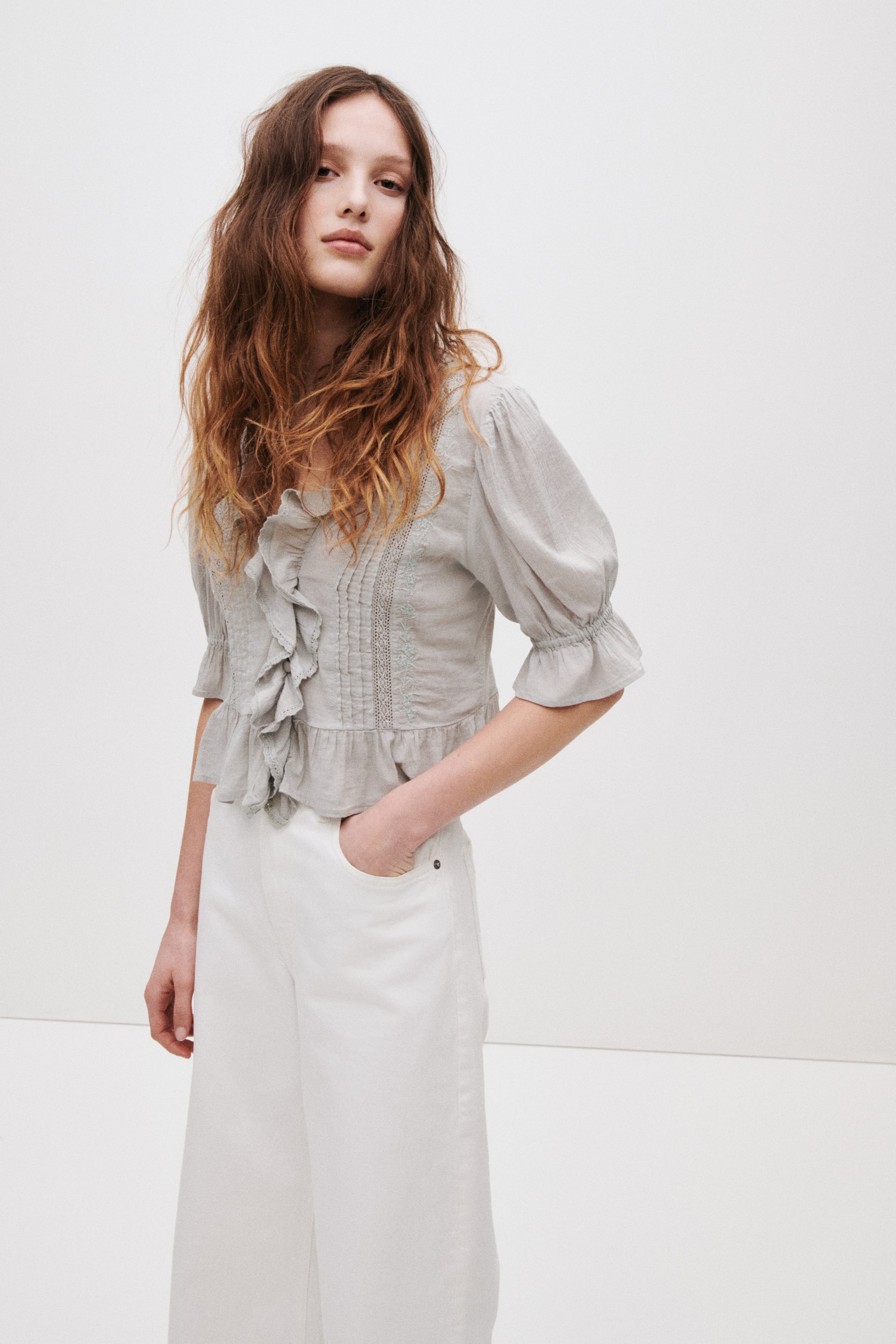 View larger image: Seorang model sedang memakai blaus tenunan kelabu muda. Ia menampilkan pintucks, trim renda, perincian bersulam, garis leher V yang dalam dengan ruffle dan butang tertutup, lengan puff panjang siku dengan elastik dan manset kembang, serta flounce lebar di hem. Model tersebut berdiri dengan satu tangan di dalam poket seluar putih, yang dipakai bersama blaus tersebut.