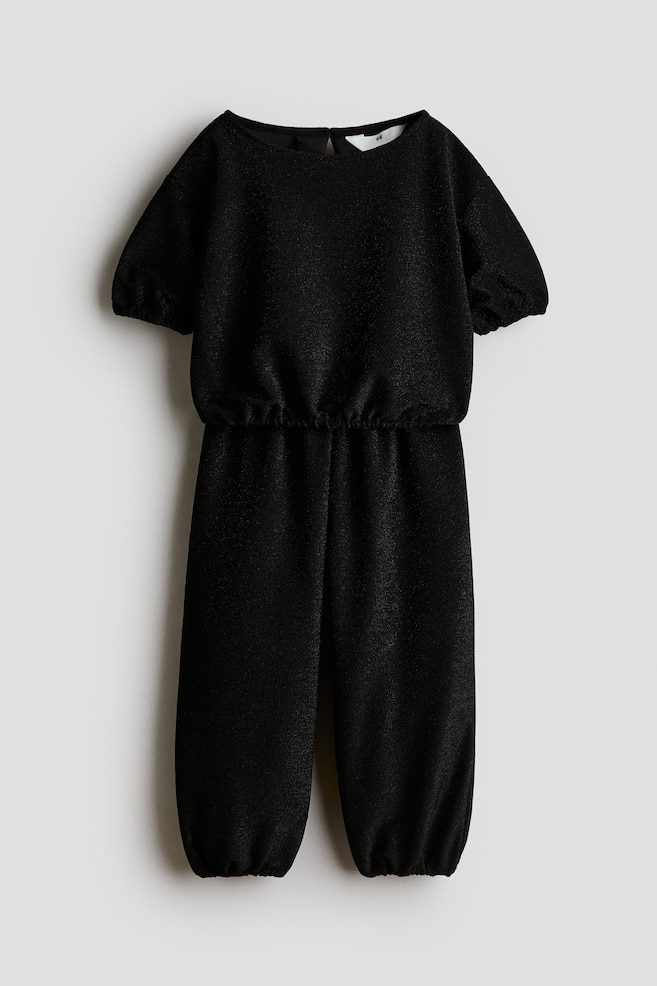 A black shimmering jumpsuit is shown front-facing, featuring short puff sleeves with elasticated cuffs, a gathered elasticated waistband, and full-length trousers with elasticated ankle hems. - 1