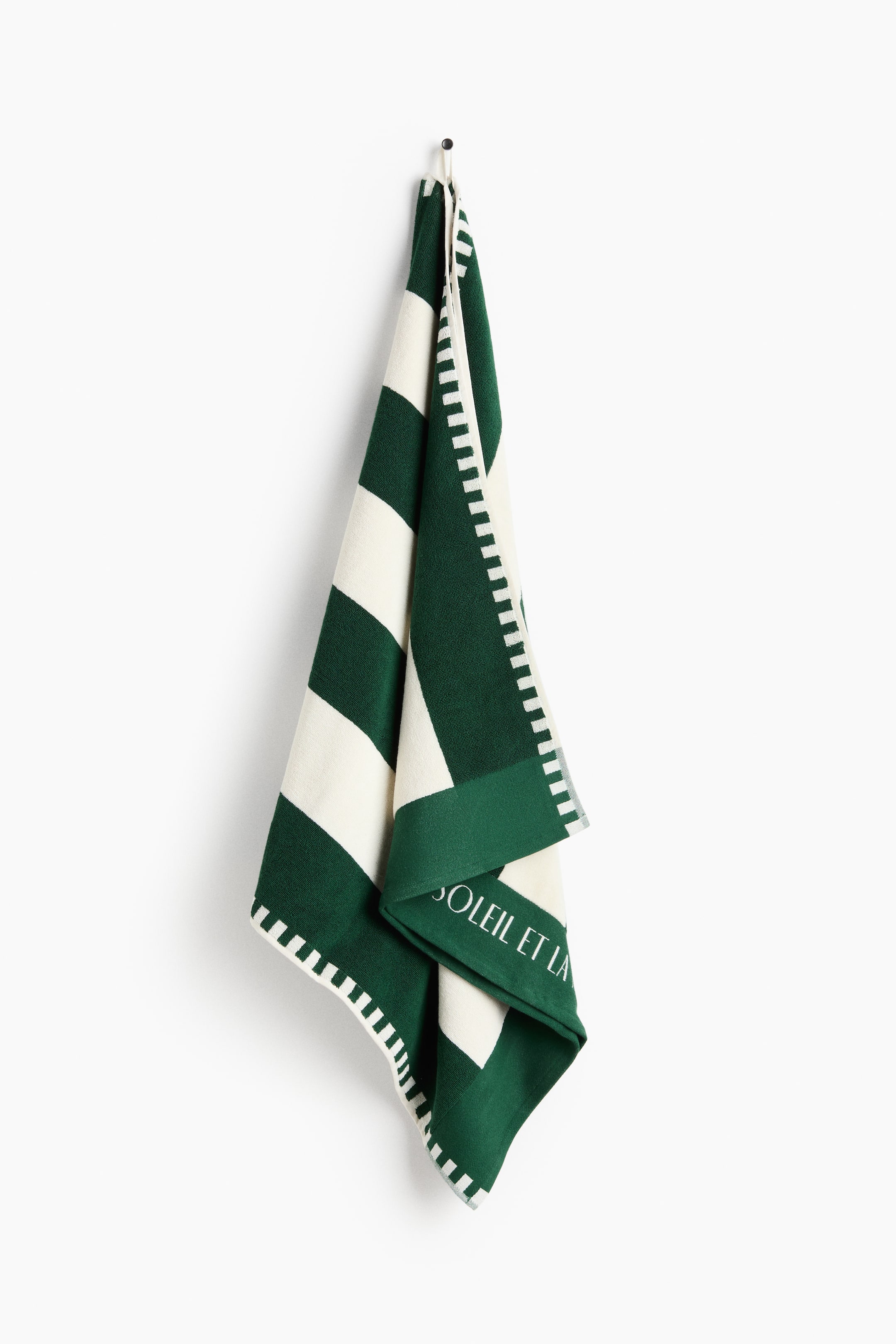 View larger image: A green and off-white striped towel is hanging, showcasing wide horizontal stripes. It features a decorative border of small white rectangles along its edges and partially visible white text, "SOLEIL ET LA," on a solid green section.