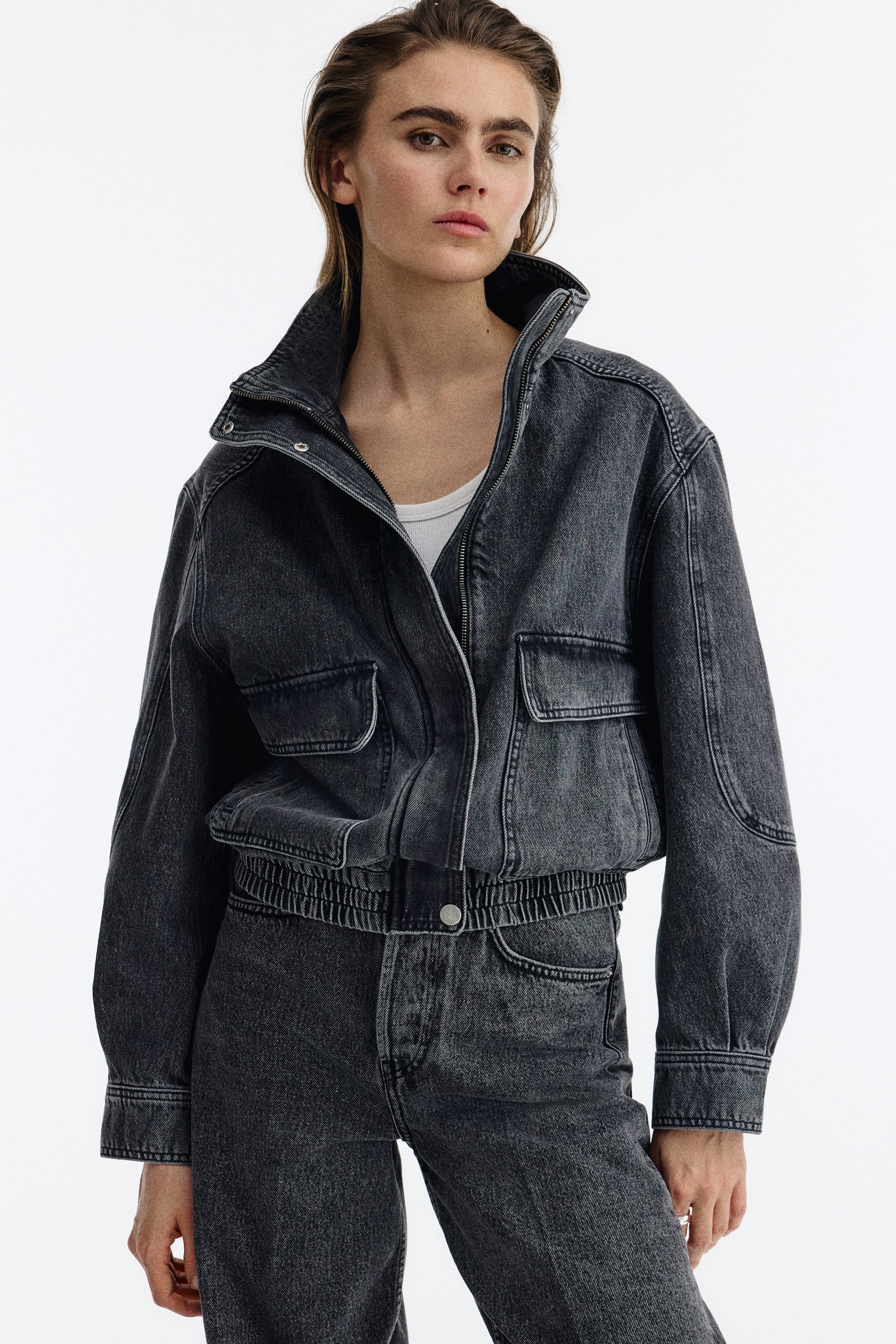 View larger image: The oversized cotton denim jacket is facing front, featuring a stand collar and a partially unzipped front with a wind flap secured by press-studs. It displays two patch front pockets with flaps, welt side pockets, press-studs at the cuffs, and smocked elastication at the hem, worn over a white vest top.