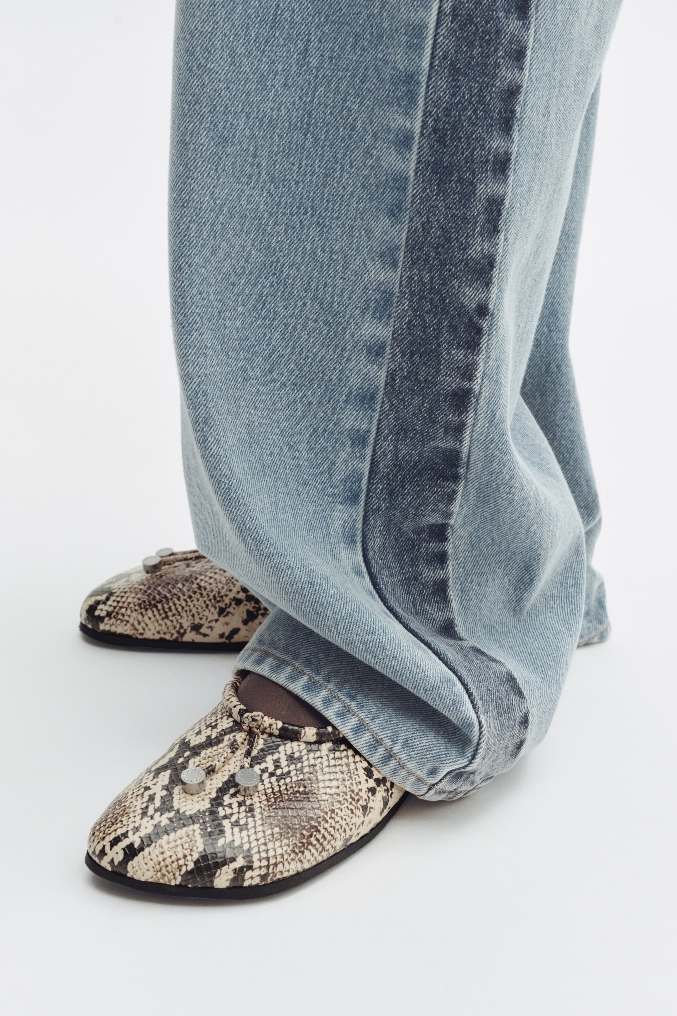 View larger image: Light blue wide-leg jeans gather around the ankles, showcasing a darker vertical stripe along the outer seam. They partially cover snakeskin print flat shoes, detailed with two silver studs on the vamp, facing forward.