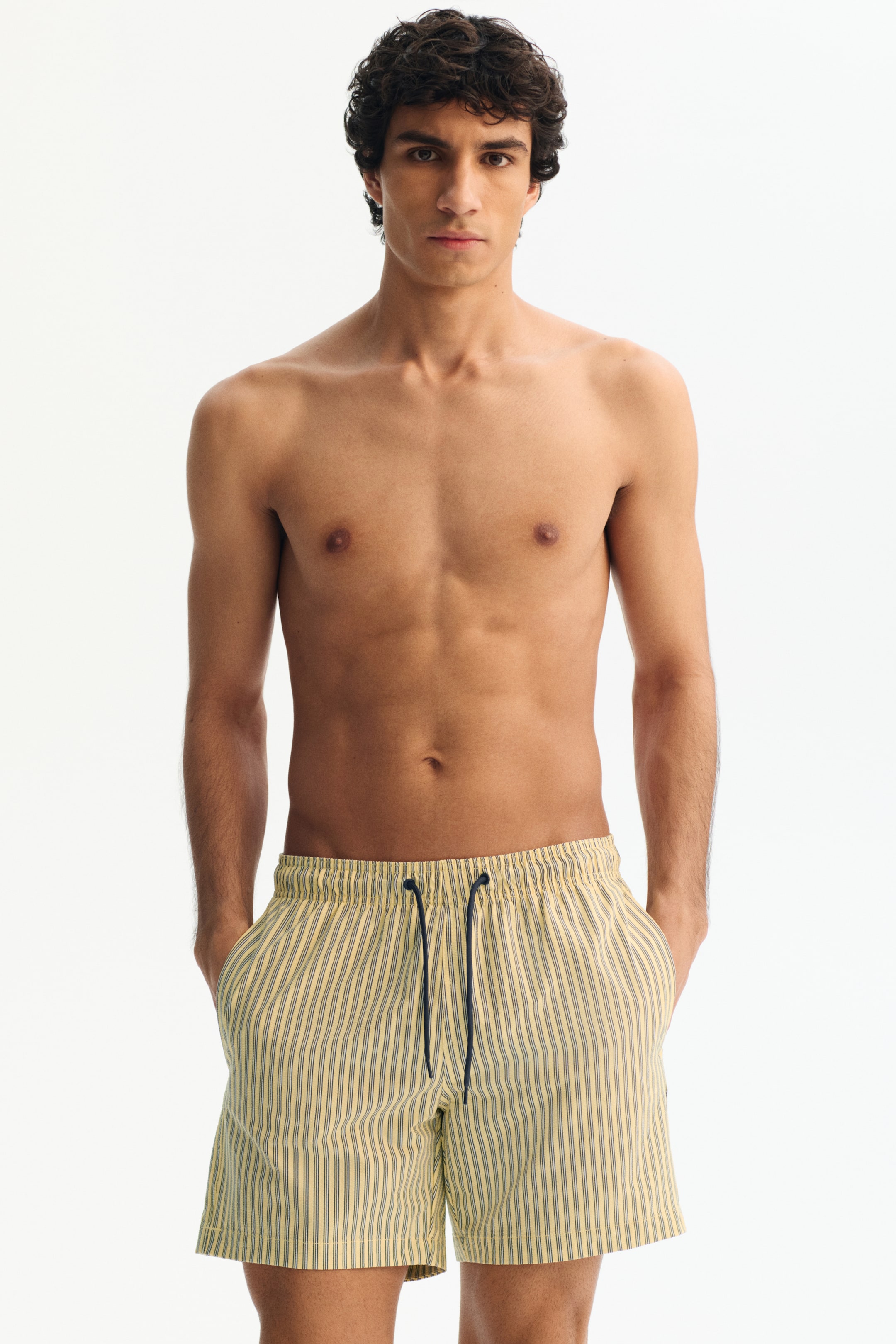 Agrandir l'image: Shorts de bain pour hommes avec un motif à rayures verticales jaunes et bleu foncé. Ils sont conçus avec une taille élastiquée et un cordon de serrage bleu foncé, avec des poches latérales visibles lorsque les mains y sont reposées.